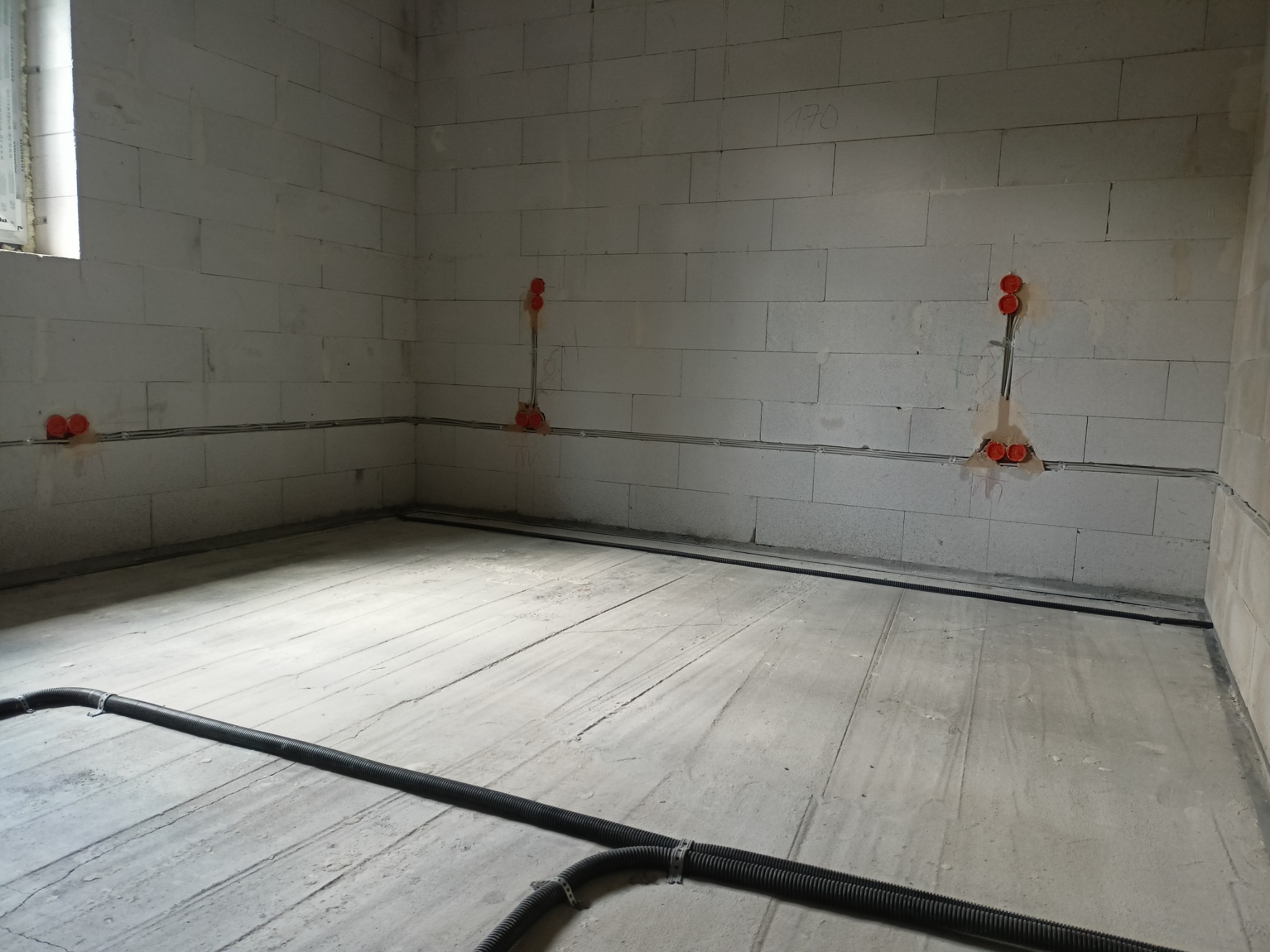 Surowy stan wnętrza z widocznymi przewodami elektrycznymi w ścianach z betonu komórkowego i rurami instalacyjnymi na podłodze.