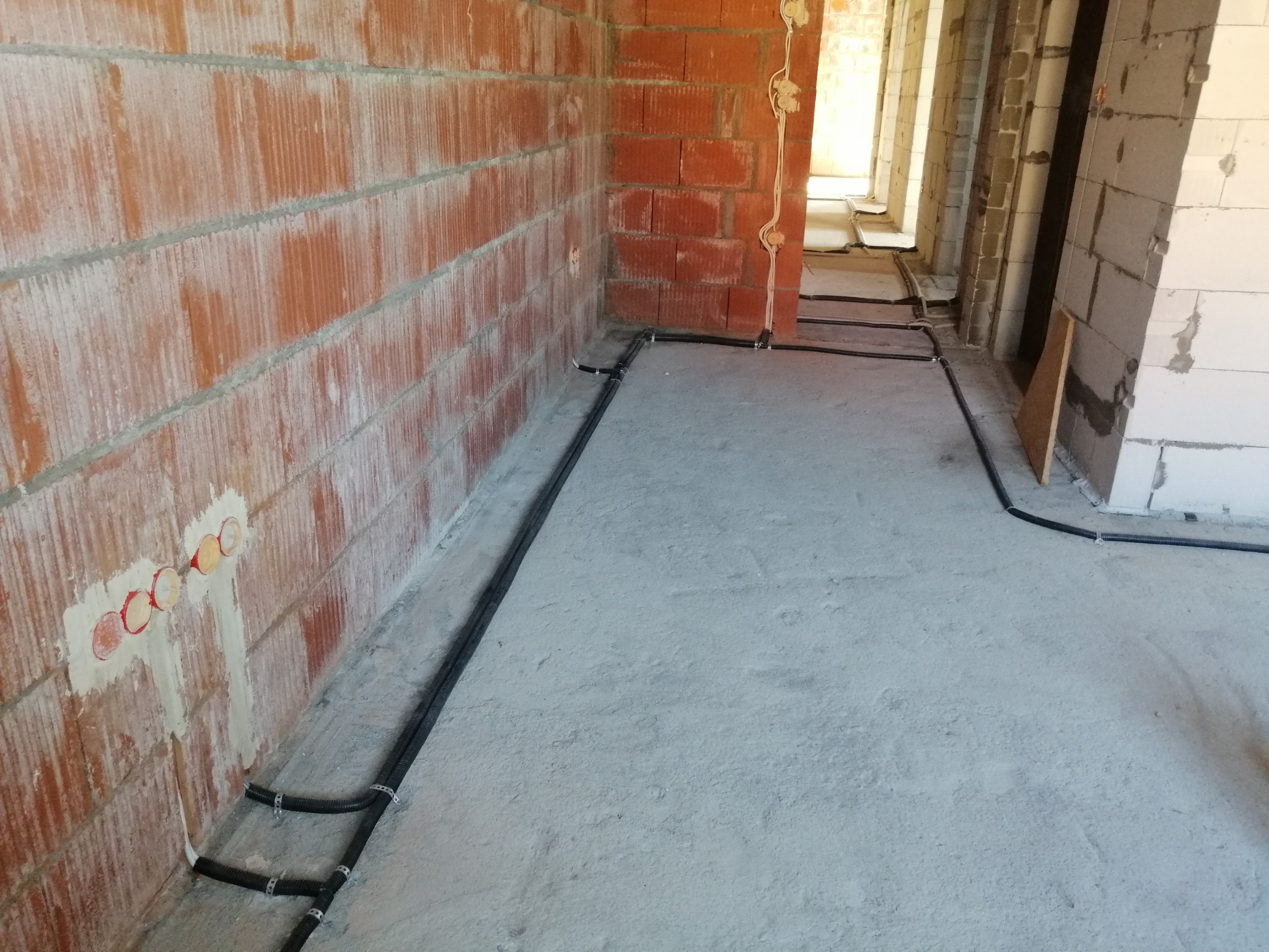 Wnętrze w trakcie prac instalacyjnych: widoczne czarne kable elektryczne prowadzone po podłodze wzdłuż ścian z czerwonej cegły i białego pustaka, z przygotowanymi otworami na gniazdka elektryczne.