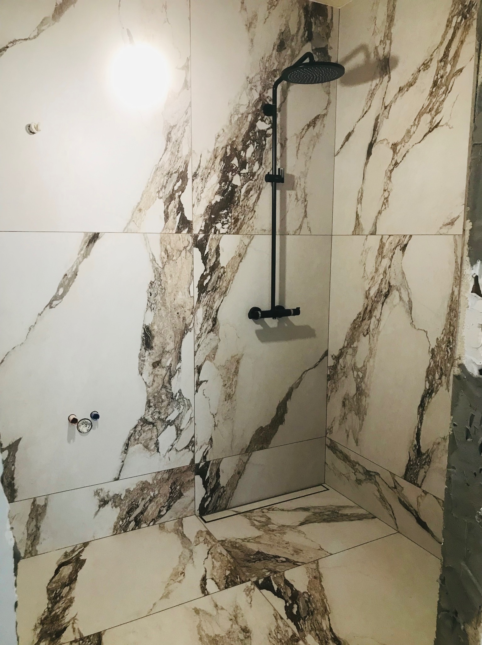 Fragment łazienki w trakcie wykańczania: marmurowe płytki na ścianach i podłodze, czarna deszczownica, widoczne elementy instalacji wodnej oraz fragment ściany z resztkami zaprawy.