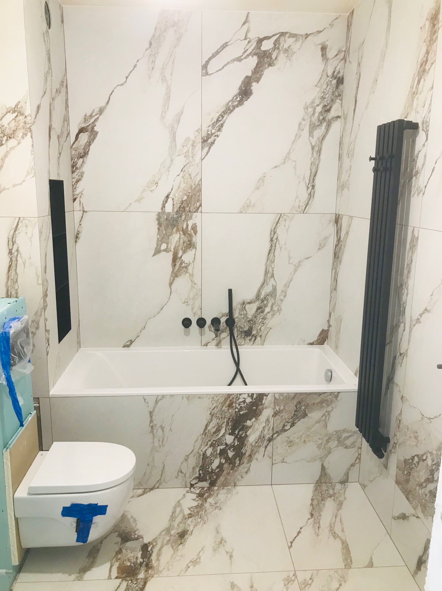 Nowoczesna łazienka w trakcie wykończenia z marmurowymi płytkami, czarną armaturą i grzejnikiem żeberkowym. Uwagę zwraca toaleta podwieszana z niebieską taśmą zabezpieczającą.