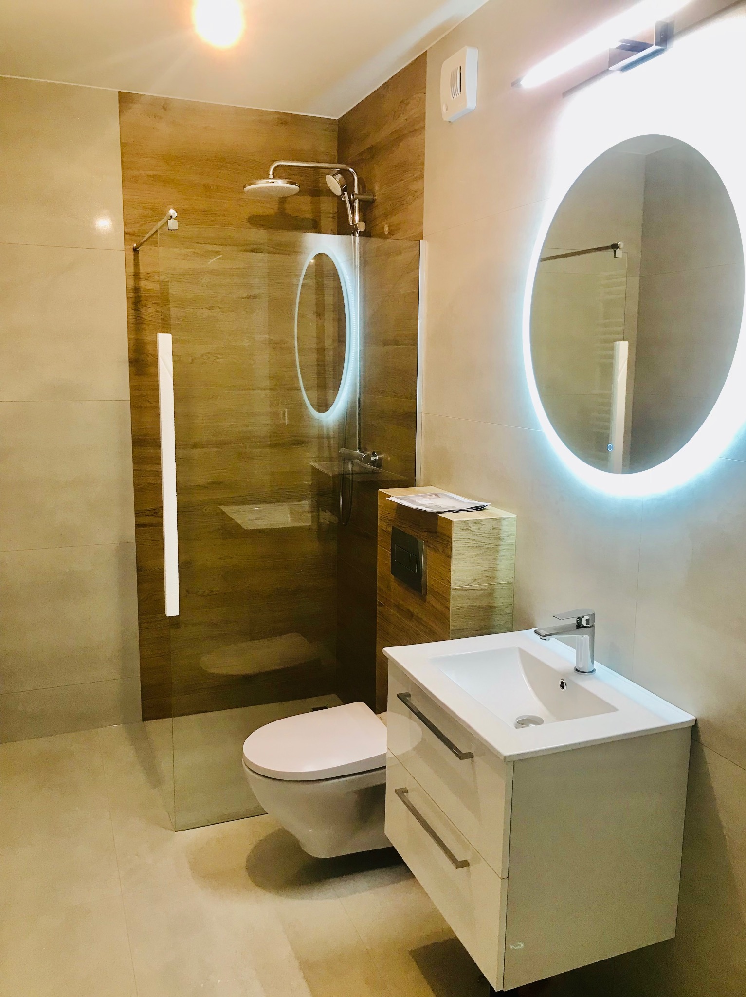 Nowoczesna łazienka z kabiną prysznicową typu walk-in, podświetlanym okrągłym lustrem, białą szafką z umywalką i podwieszaną toaletą; ściany wyłożone płytkami imitującymi drewno i beżowymi.
