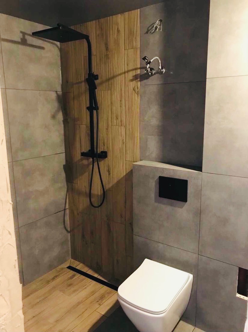 Nowoczesna łazienka z kabiną prysznicową typu walk-in, wykończona szarymi płytkami i drewnopodobnymi panelami, z czarną armaturą i odpływem liniowym.