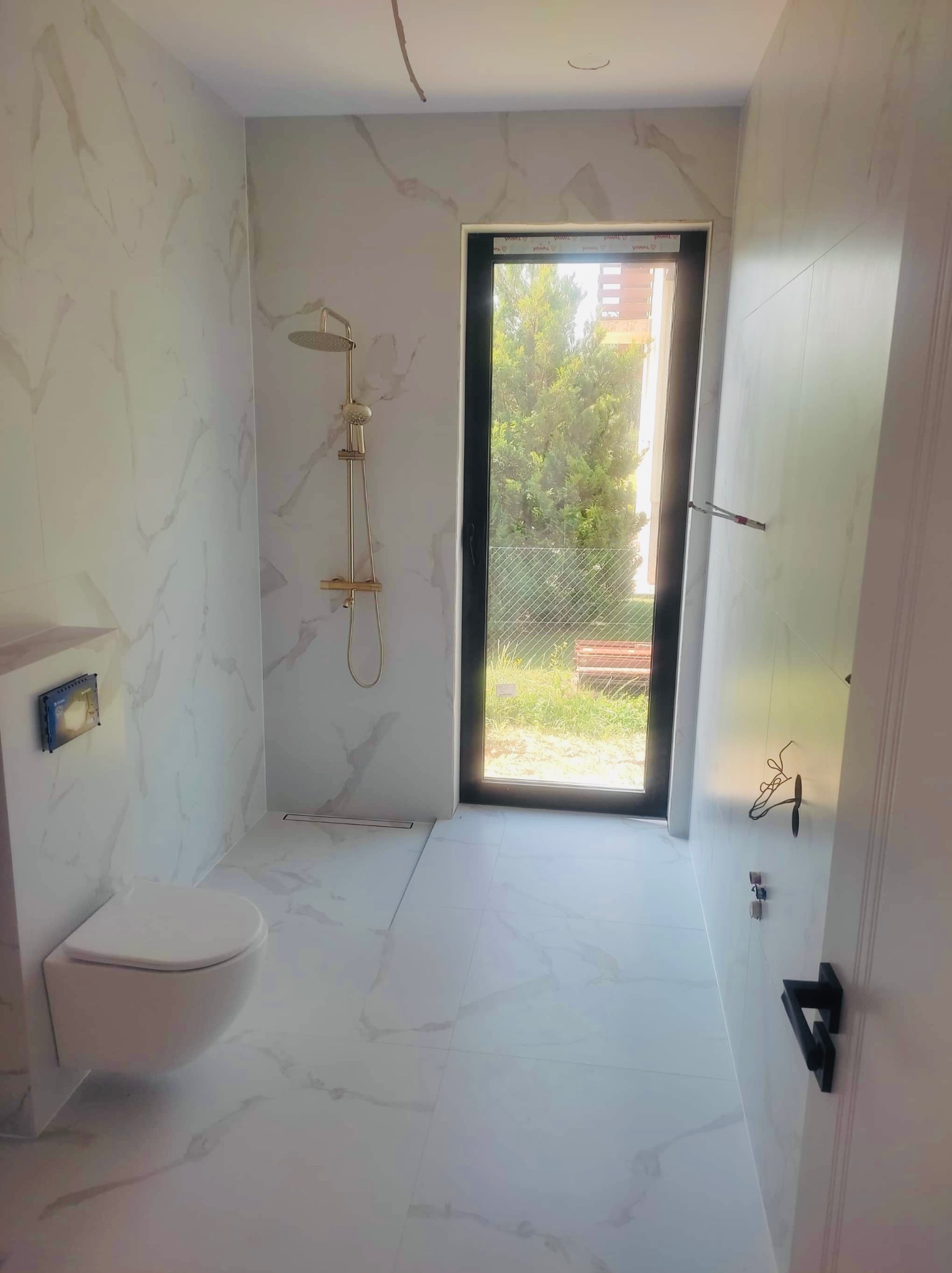 Nowoczesna łazienka z białymi płytkami imitującymi marmur, wisząca toaleta, złota armatura prysznicowa i odpływ liniowy, czarna rama okienna z widokiem na zieleń.