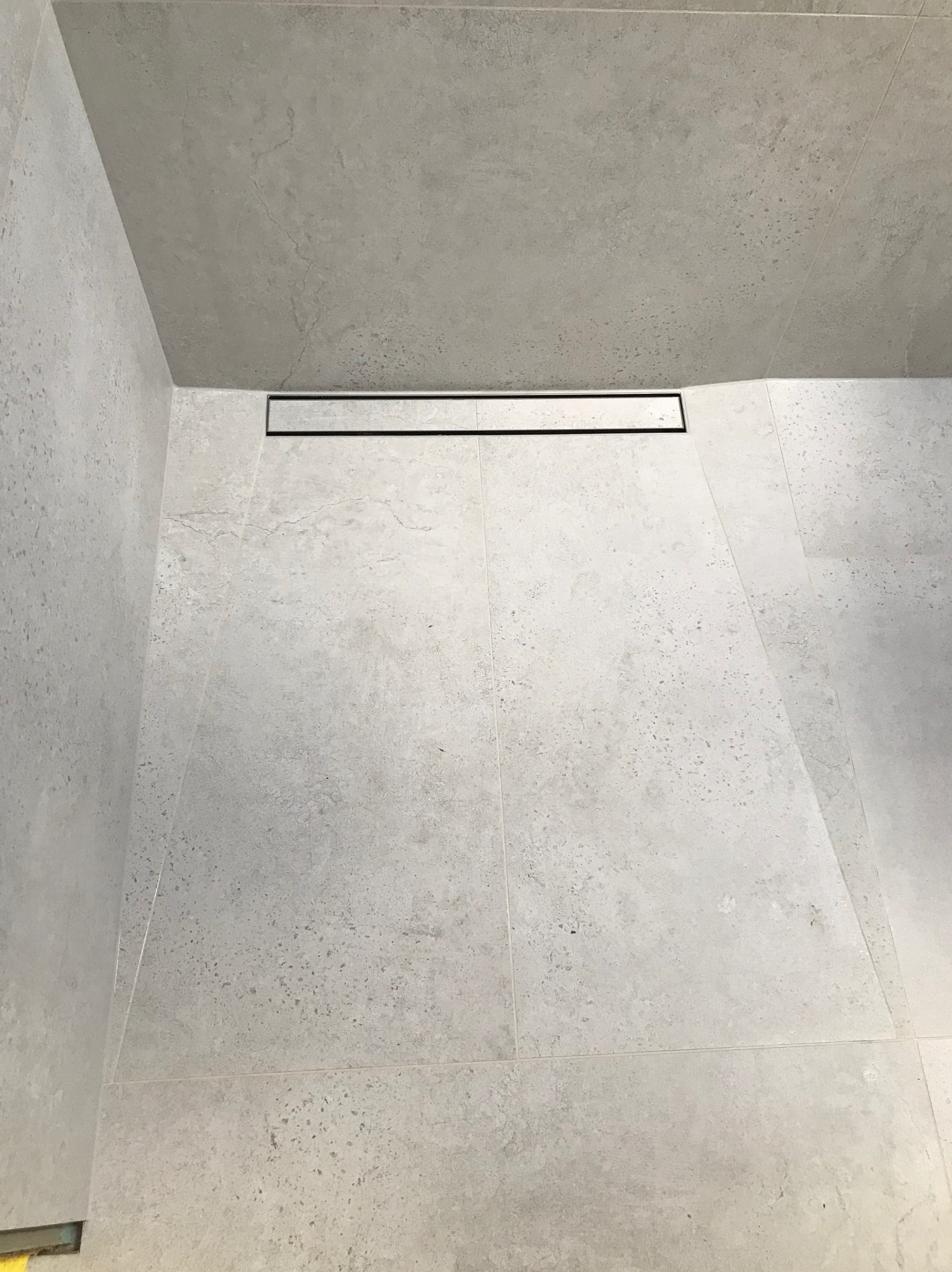 Szare płytki podłogowe i ścienne w kabinie prysznicowej z odpływem liniowym w kolorze czarnym.