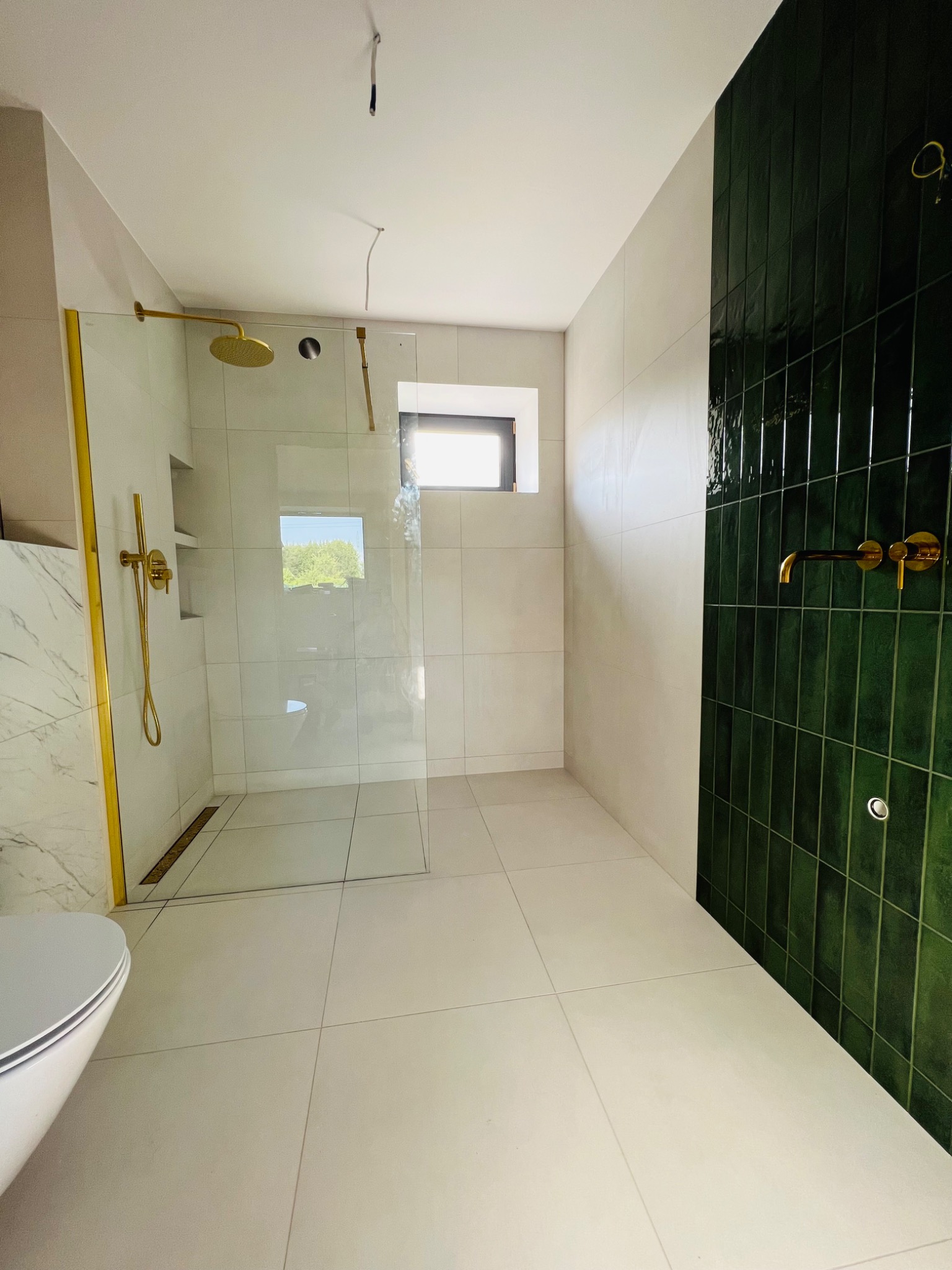 Nowoczesna łazienka z kabiną prysznicową ze złotymi ramami, zielonymi płytkami na ścianie i oknem.