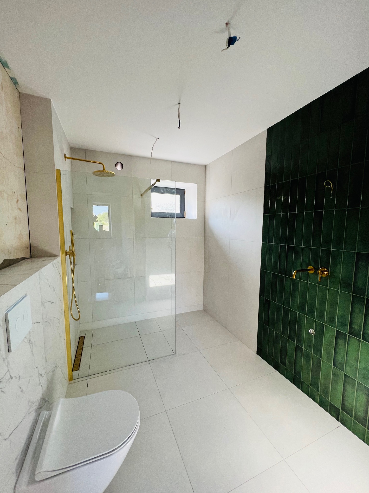 Nowoczesna łazienka w trakcie wykończenia: kabina prysznicowa ze złotymi elementami, pionowa ściana z ciemnozielonych płytek, biała toaleta i widoczne niedokończone prace na ścianie.