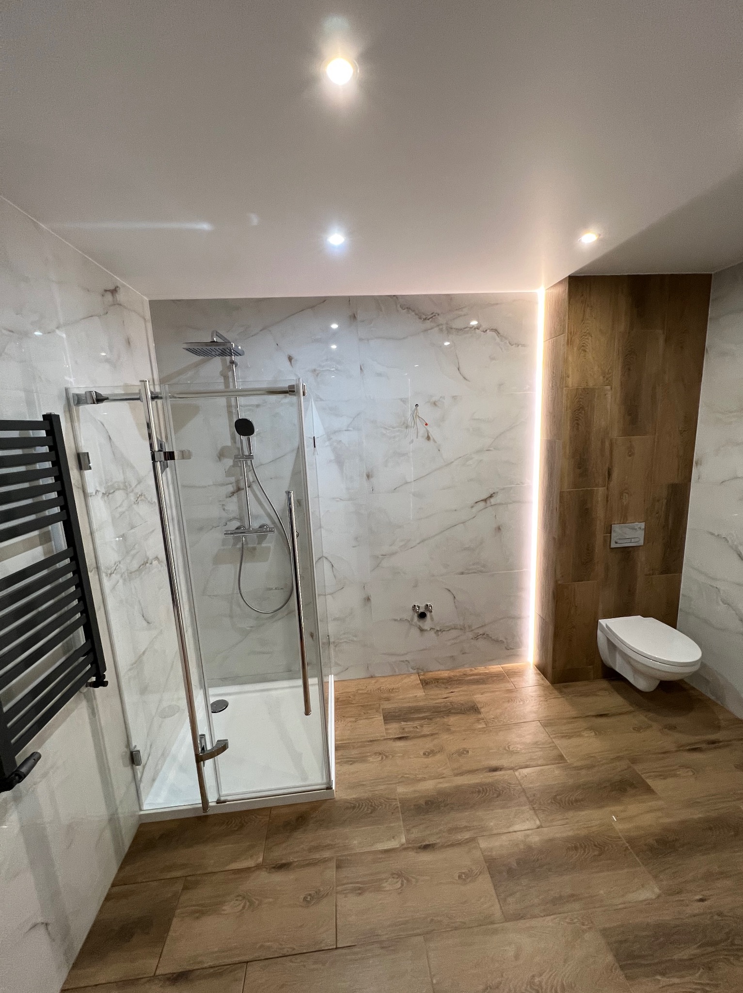 Nowoczesna łazienka z kabiną prysznicową typu walk-in, marmurowymi płytkami na ścianach i drewnopodobnymi na podłodze. Oświetlenie LED wzdłuż ściany dodaje elegancji.