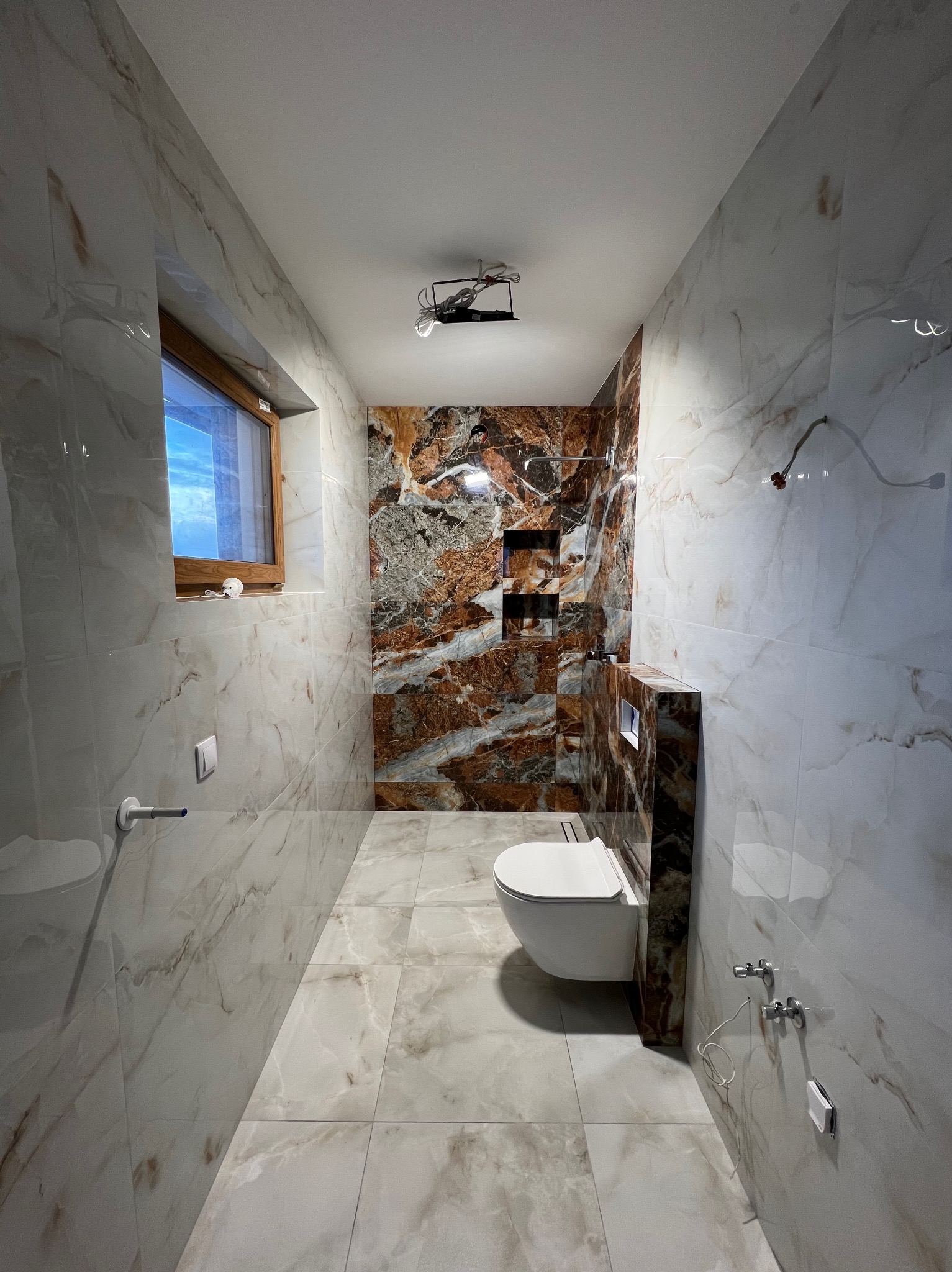 Wąska łazienka z marmurowymi płytkami w odcieniach bieli i brązu. Widoczna podwieszana toaleta, okno i instalacja oświetleniowa w trakcie montażu.