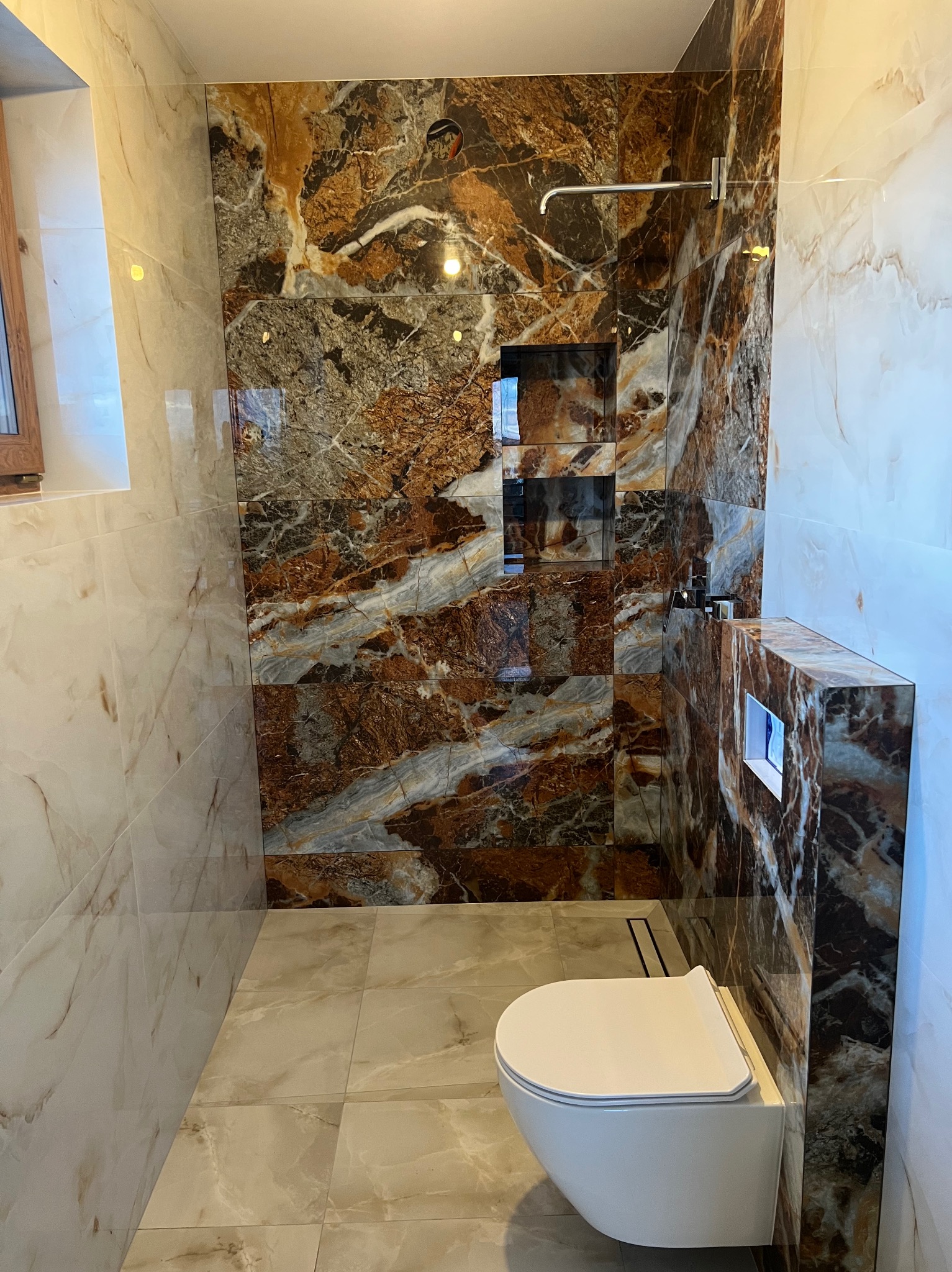 Nowoczesna łazienka z kabiną prysznicową typu walk-in, wykończona płytkami imitującymi marmur w odcieniach brązu i beżu. Widoczny podwieszany sedes i wnęki w ścianie.