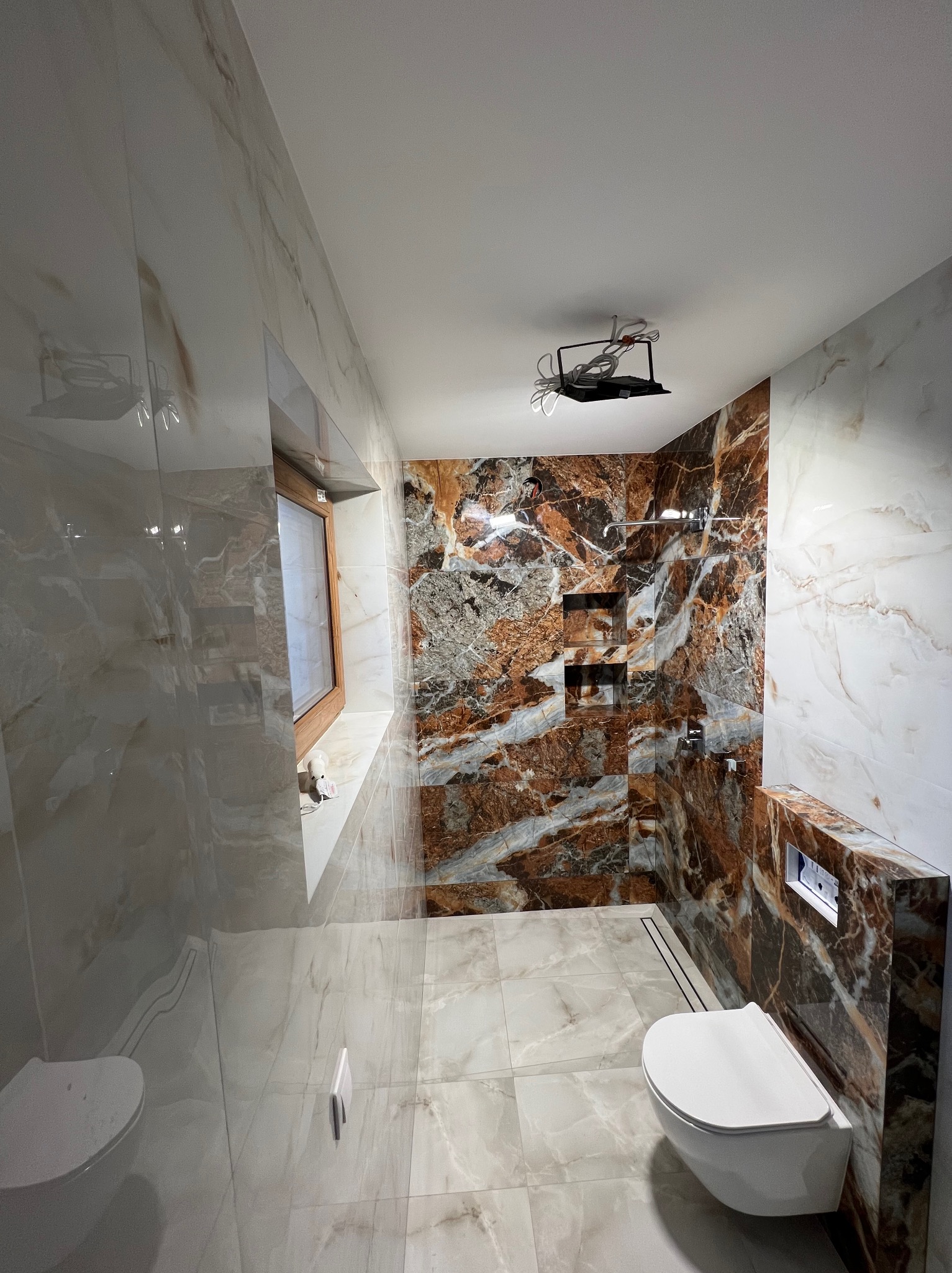 Wykończona łazienka z marmurowymi płytkami w odcieniach beżu i brązu. Kabina prysznicowa typu walk-in z odpływem liniowym. Podwieszana toaleta.