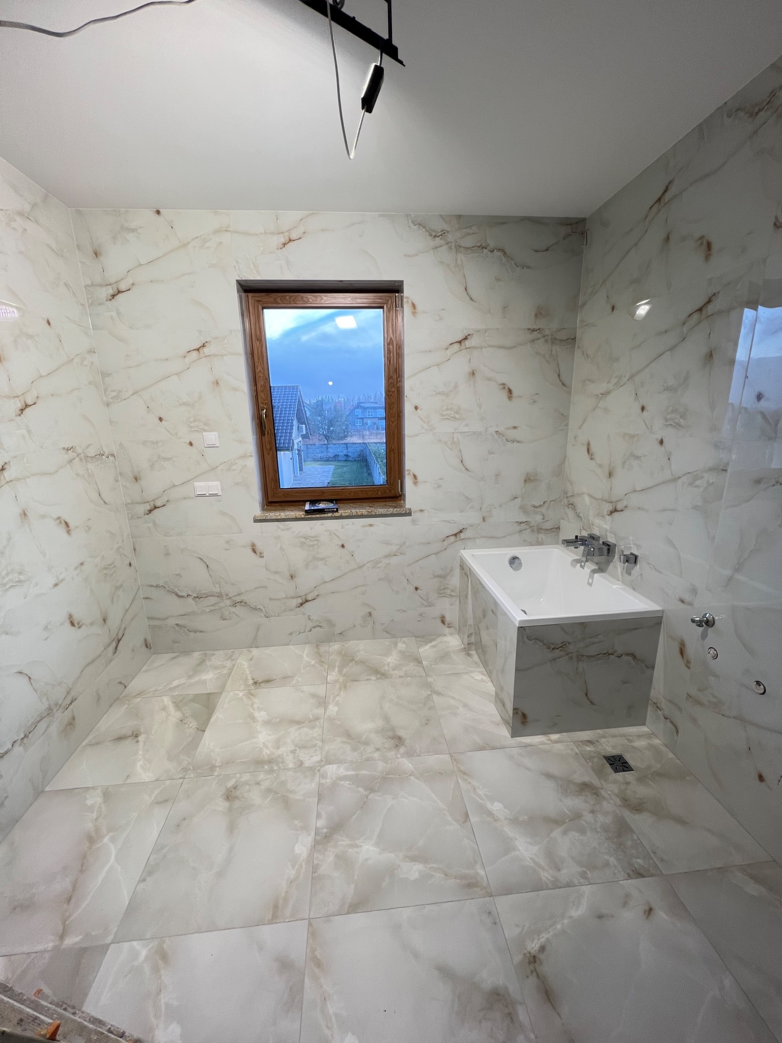 Wykończona łazienka z wanną obudowaną płytkami imitującymi marmur. Widoczne okno z drewnianą ramą i fragment zabudowy prysznicowej. Nowoczesny design.