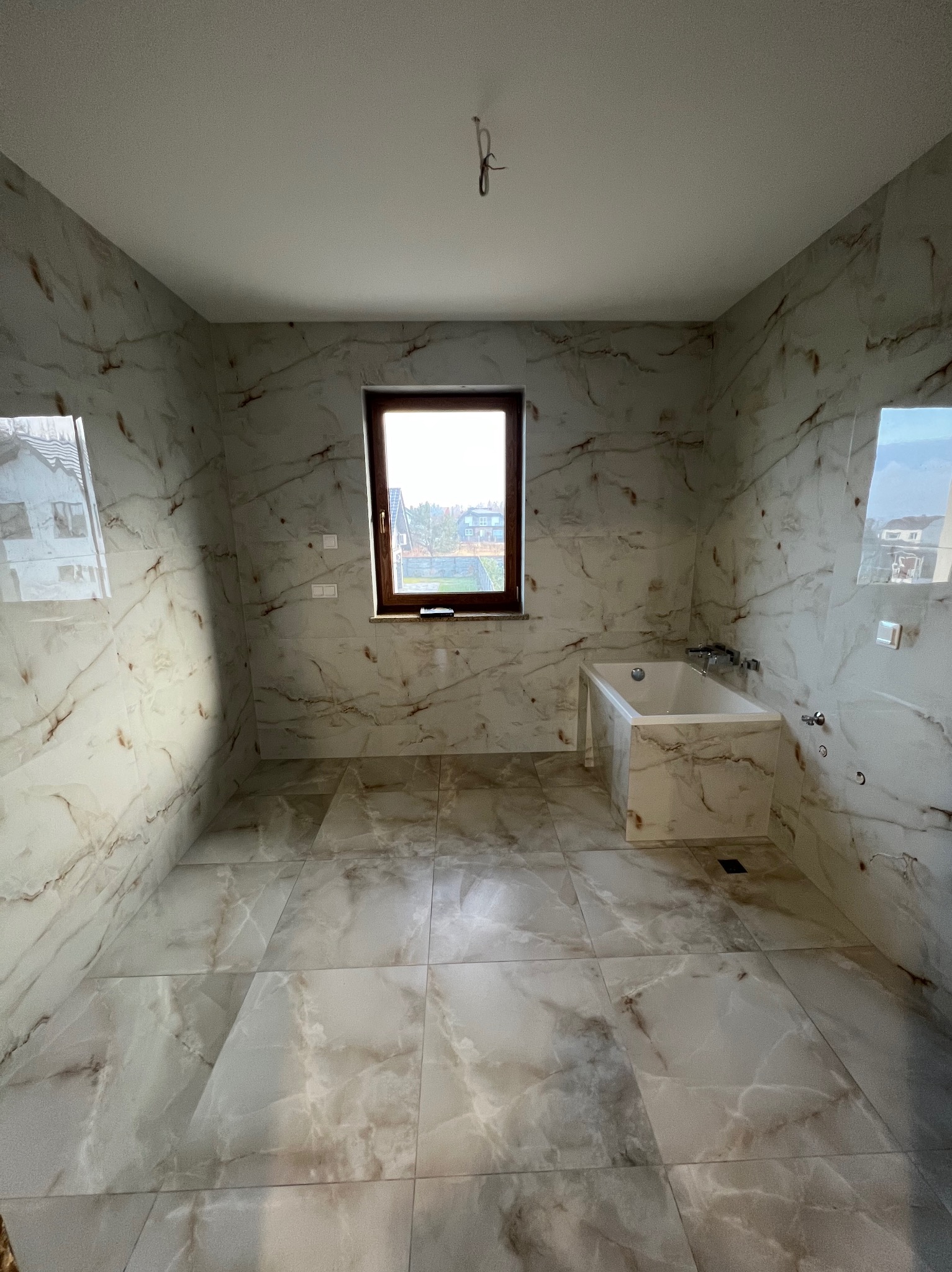 Wykończona łazienka z wanną obudowaną płytkami imitującymi marmur, okno z brązową ramą, płytki na podłodze i ścianach w odcieniach bieli i beżu.