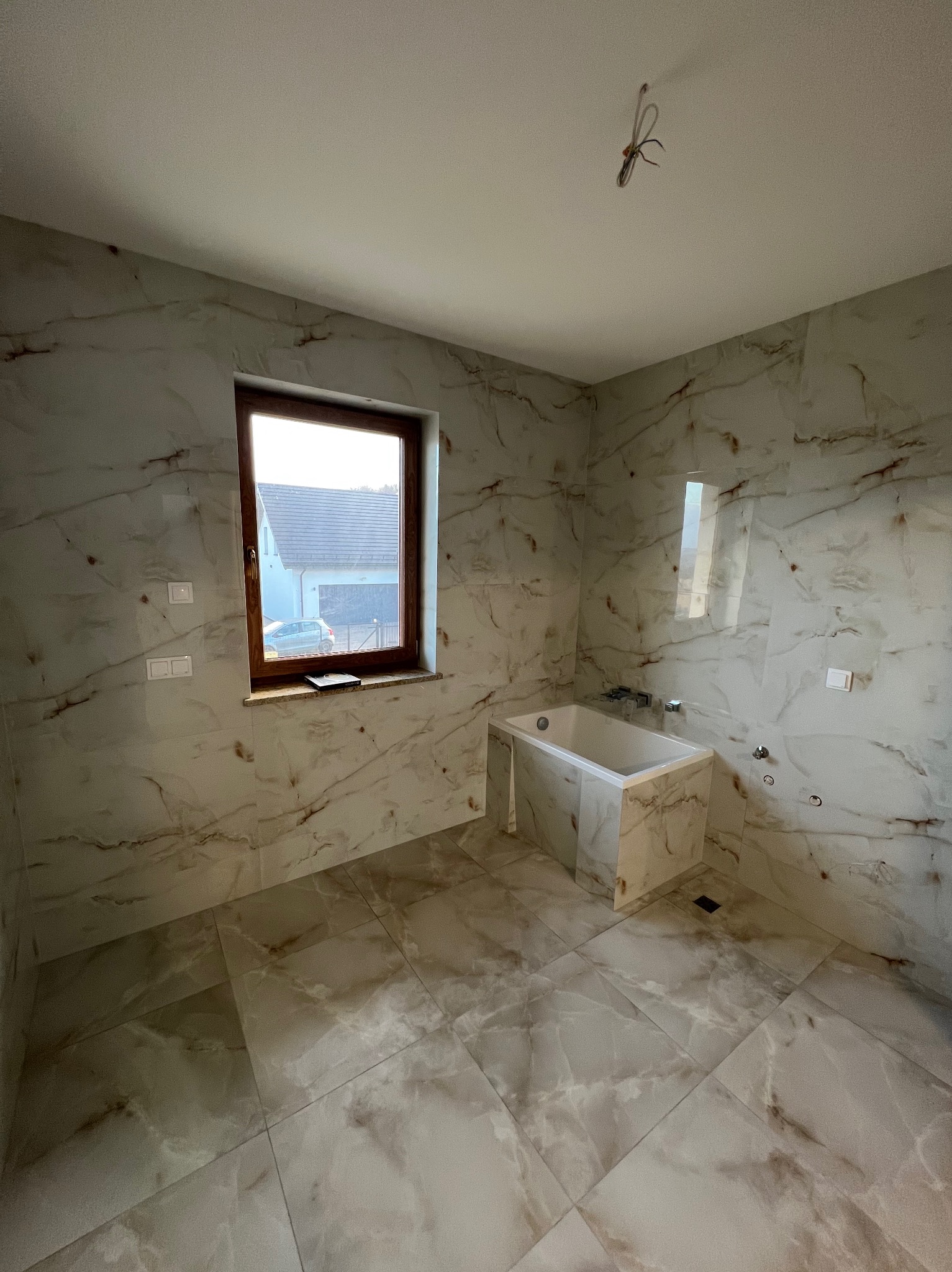 Wykończona łazienka z wanną obudowaną płytkami imitującymi marmur. Widoczne okno i wystające kable ze sufitu.