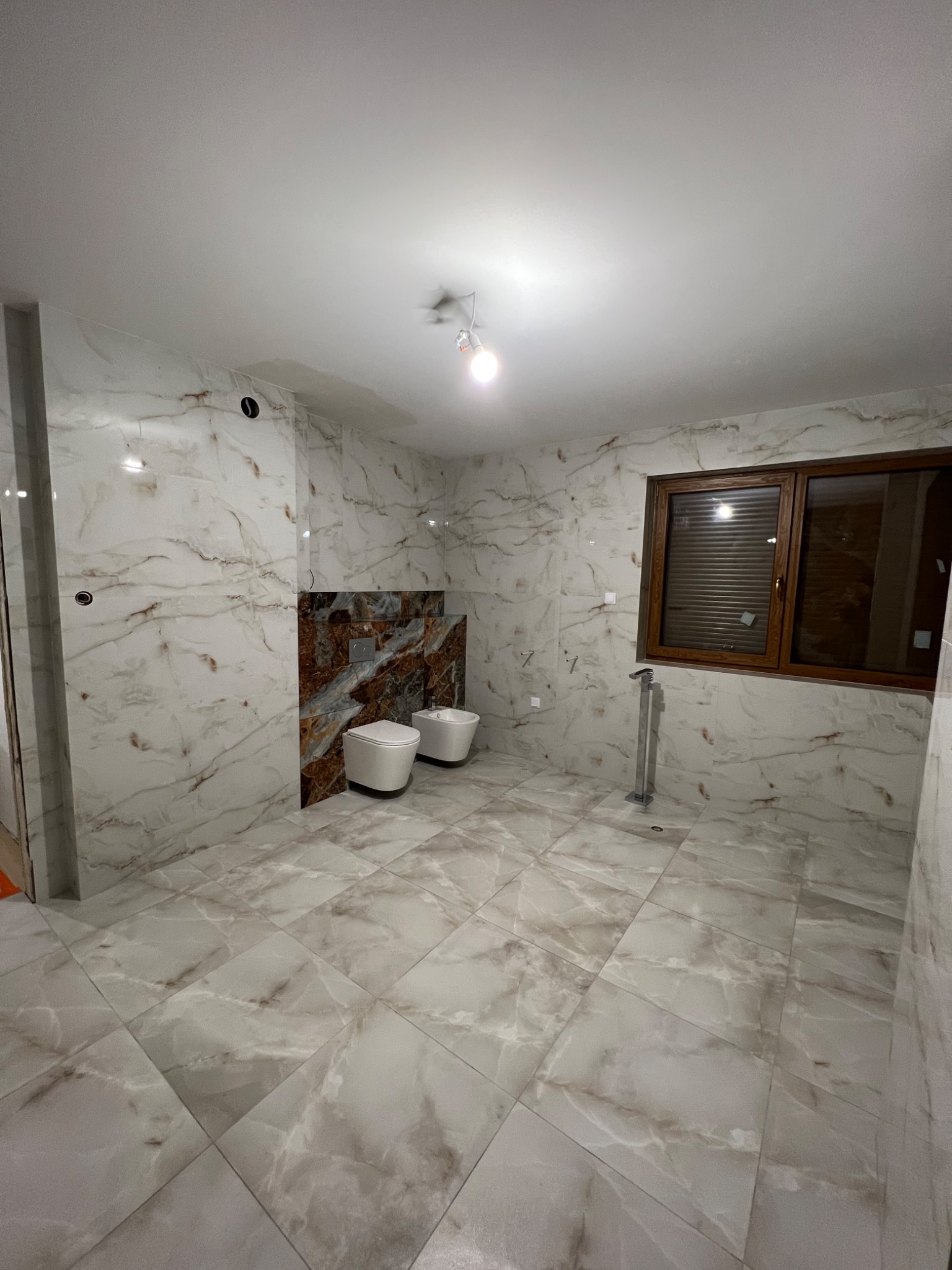 Wykończona łazienka z marmurowymi płytkami, brązowymi akcentami i nowoczesną armaturą. Widoczny bidet i toaleta. Okno z brązowymi żaluzjami.