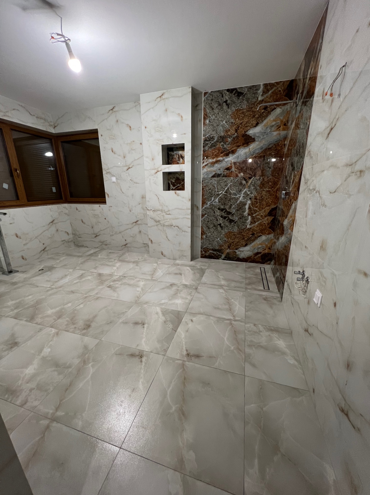 Wykończona łazienka z marmurowymi płytkami, brązowe okno i prysznic z ciemnymi kaflami. Minimalistyczny design, surowe oświetlenie.