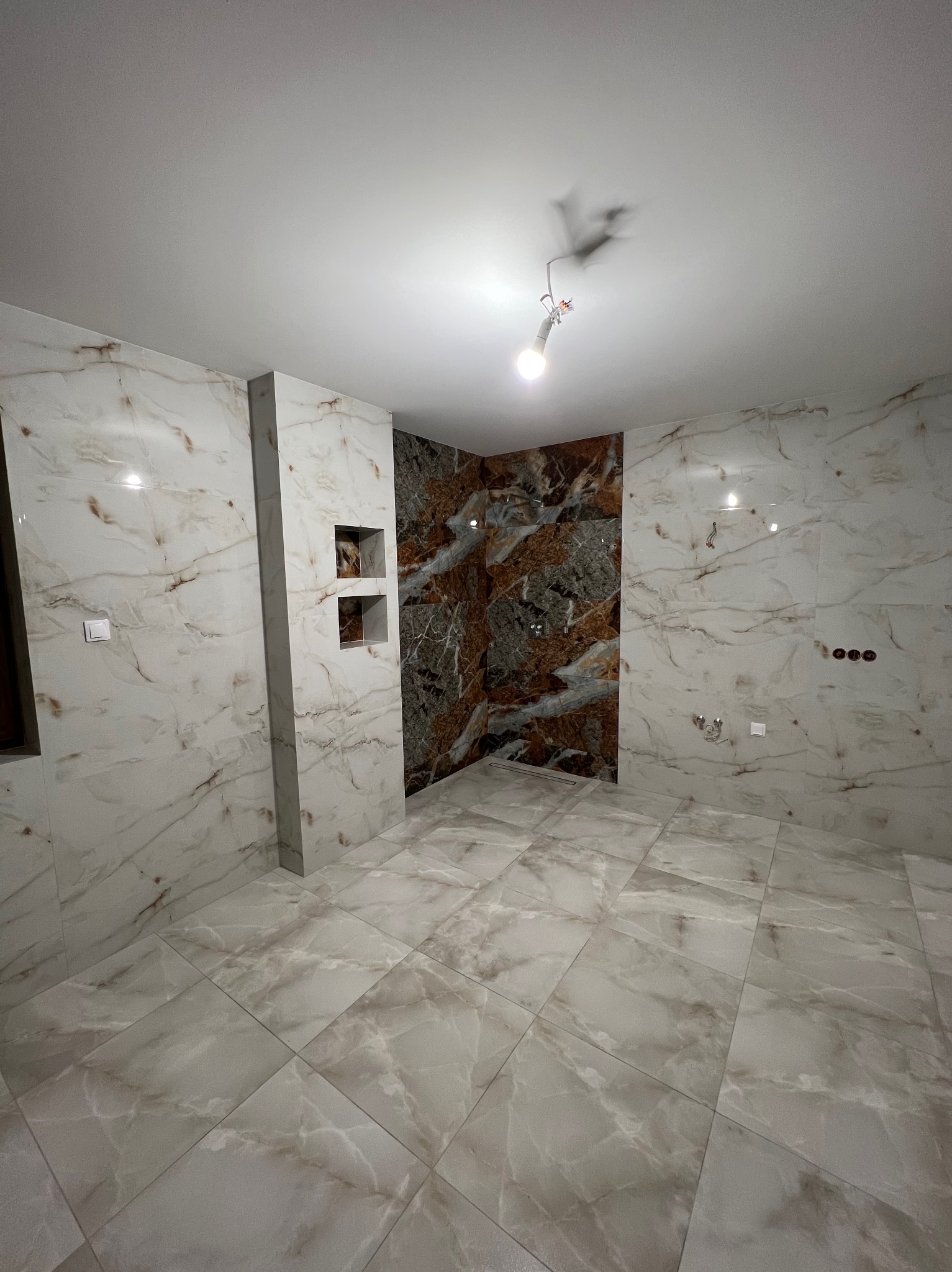 Wykończona łazienka z marmurowymi płytkami na ścianach i podłodze. Widoczne wnęki w ścianie i przygotowane punkty elektryczne. Centralnie umieszczona żarówka.