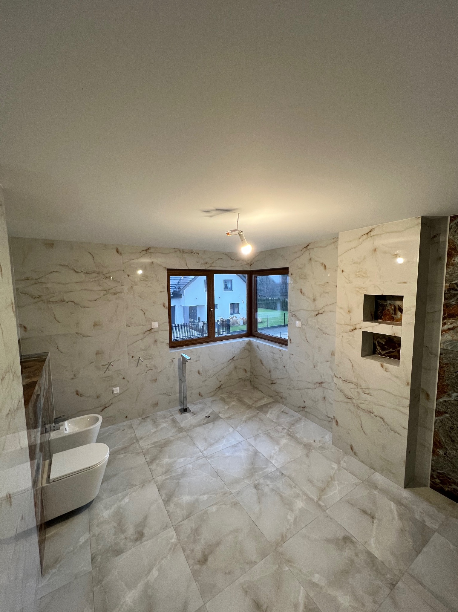 Wykończona łazienka z marmurowymi płytkami, wiszącą toaletą i bidetem. Widoczne okno z widokiem na dom i ogród. Oświetlenie sufitowe.