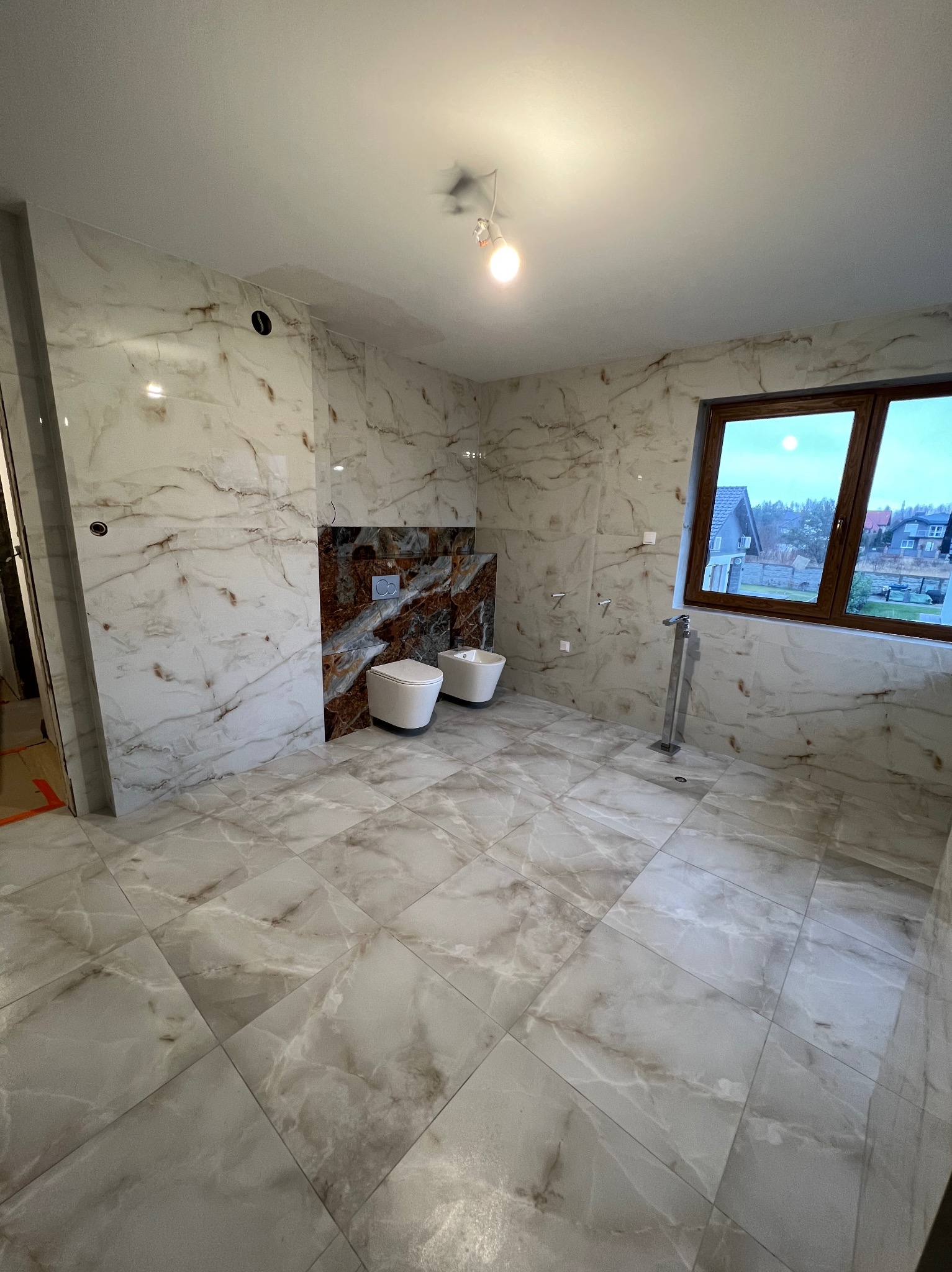 Wykończona łazienka z marmurowymi płytkami na ścianach i podłodze. Umywalka i bidet podwieszane na ścianie z brązowymi płytkami. Okno z widokiem na domy.