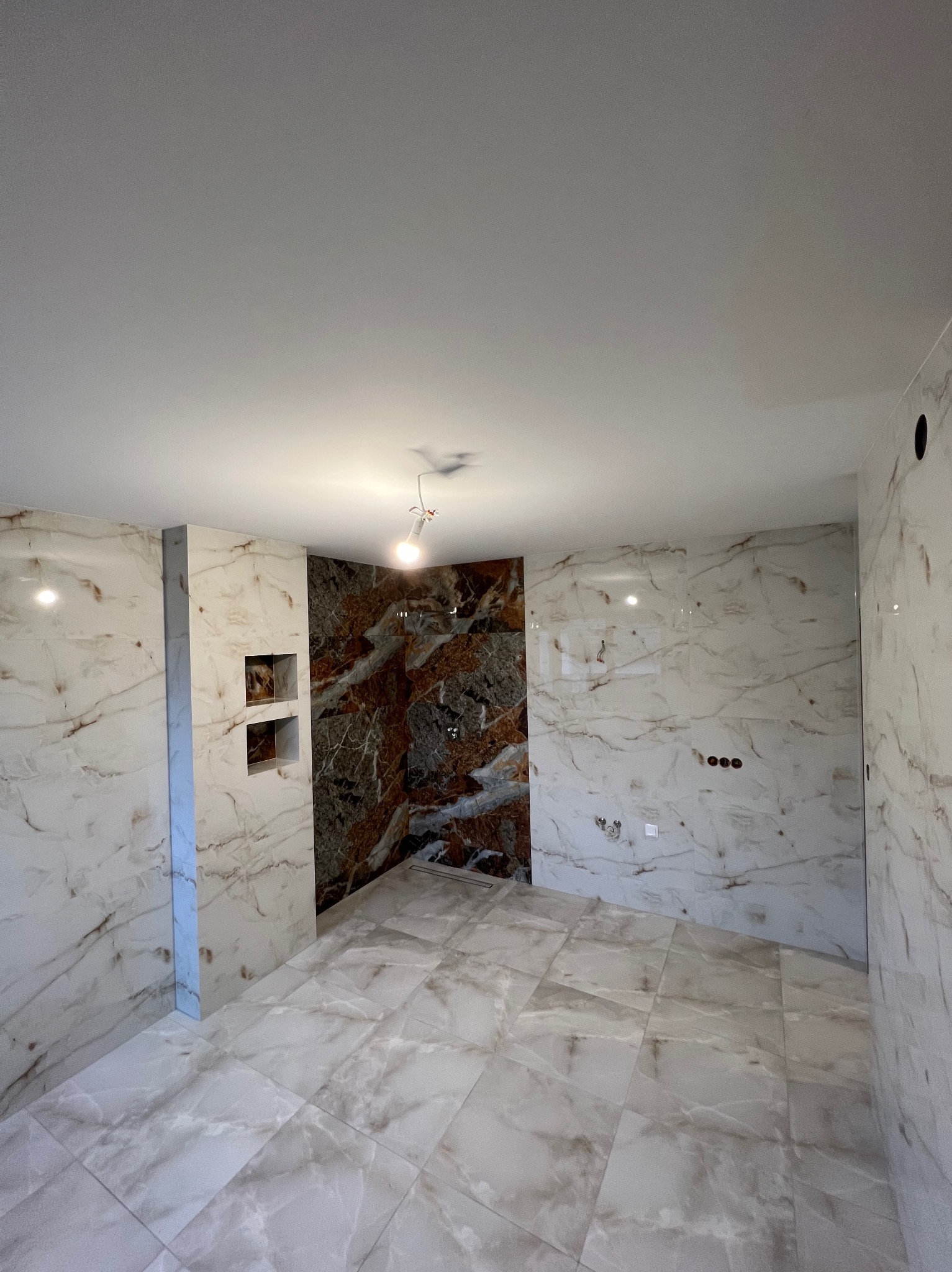 Wykończona łazienka z marmurowymi płytkami w odcieniach bieli i brązu. Widoczne wnęki ścienne i podłoga wyłożona płytkami. Oświetlenie sufitowe.