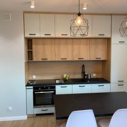 MICHAŁ WIĘCKOWSKI - PROJEKTOWANIE WNĘTRZ - Nowoczesna kuchnia z jasnymi szafkami górnymi i drewnopodobnymi dolnymi, czarnym blatem i geometrycznymi lampami wiszącymi nad stołem.