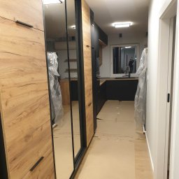 MICHAŁ WIĘCKOWSKI - PROJEKTOWANIE WNĘTRZ - Korytarz w trakcie wykańczania z zabudowaną szafą z lustrami i drewnianymi frontami, widok na czarną kuchnię z oknem.