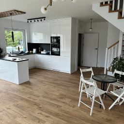 MICHAŁ WIĘCKOWSKI - PROJEKTOWANIE WNĘTRZ - Jasna kuchnia z białymi, błyszczącymi szafkami i czarnym blatem w Gdańsku, widok na stół z krzesłami i schody z drewnianymi stopniami.