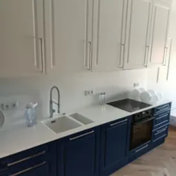 Nowoczesna kuchnia z białymi szafkami górnymi i granatowymi dolnymi, biały blat z marmurowym wzorem, zlewozmywak dwukomorowy i płyta indukcyjna, drewniana podłoga ułożona w jodełkę.