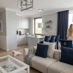 MICHAŁ WIĘCKOWSKI - PROJEKTOWANIE WNĘTRZ - Jasne wnętrze z otwartą kuchnią i salonem. Białe meble, drewniana podłoga, niebieskie akcenty. Kobieta stoi przy stole, a mały pies leży na podłodze.