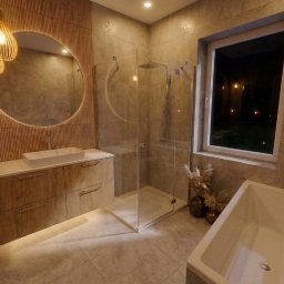 MICHAŁ WIĘCKOWSKI - PROJEKTOWANIE WNĘTRZ - Nowoczesna łazienka z okrągłym lustrem podświetlonym LED, kabiną prysznicową i wanną. Umywalka nablatowa na szafce z drewnianymi frontami. Ciepłe oświetlenie.