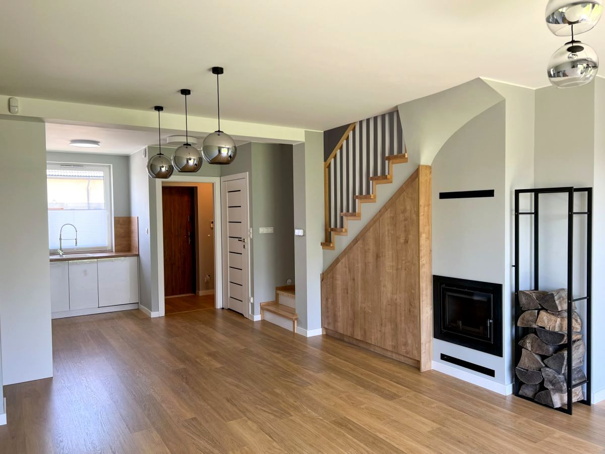 Jasne wnętrze domu z drewnianą podłogą, kominkiem z czarną obudową, schodami z drewnianą balustradą w biało-szare paski i trzema wiszącymi lampami kulistymi.