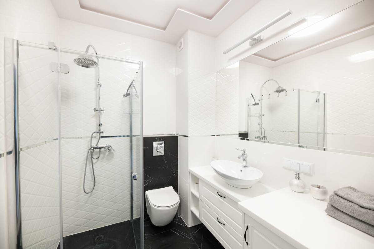 Nowoczesna, jasna łazienka z kabiną prysznicową ze szklanymi drzwiami, umywalką nablatową na białej szafce z szufladami, czarno-białymi płytkami ściennymi z geometrycznym wzorem i czarną podłogą...