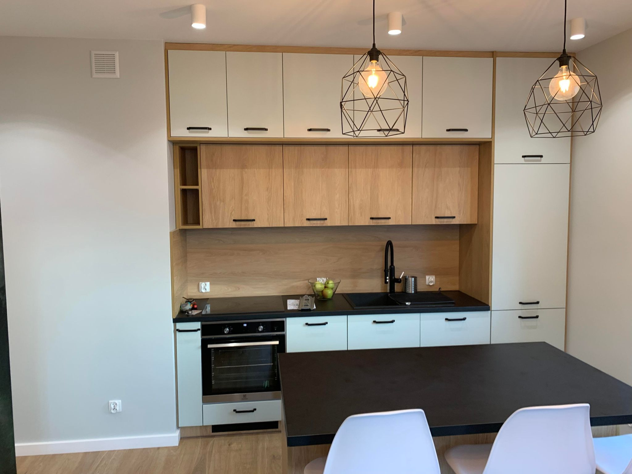 Nowoczesna kuchnia z jasnymi szafkami górnymi i drewnopodobnymi dolnymi, czarnym blatem i geometrycznymi lampami wiszącymi nad stołem.