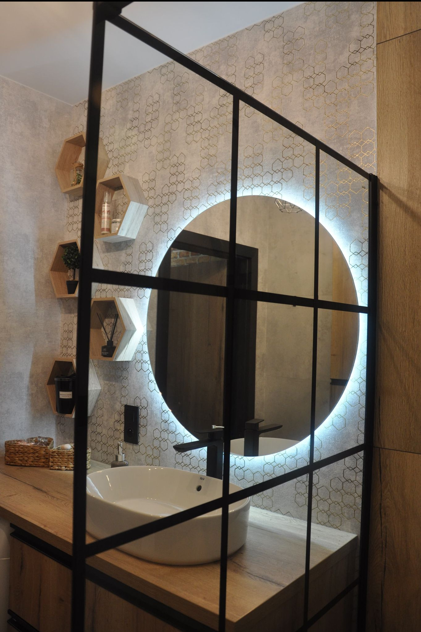 Nowoczesna łazienka z okrągłym lustrem podświetlanym LED, umywalką nablatową na drewnianym blacie, heksagonalnymi półkami i ścianą w geometryczny wzór.