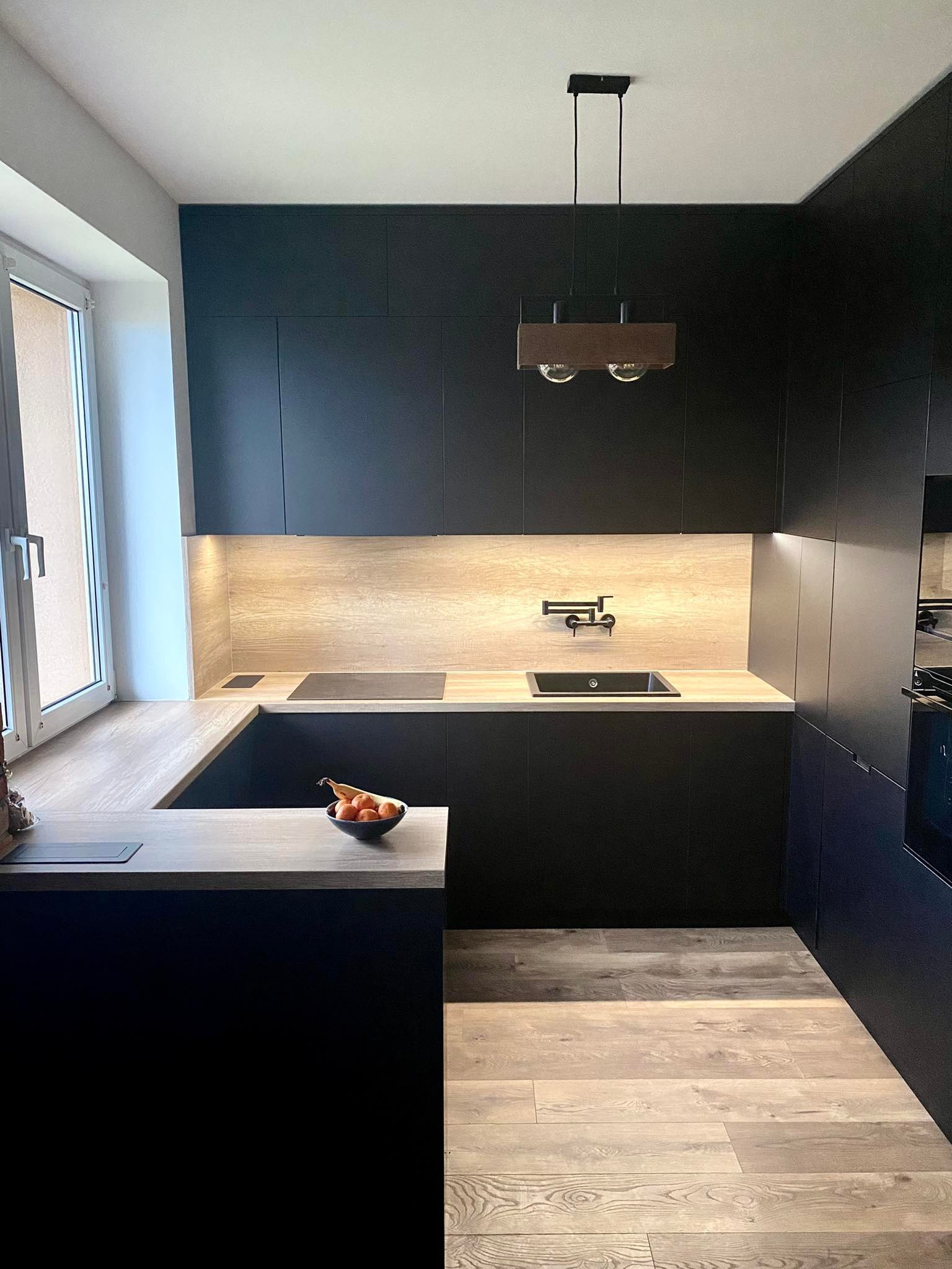 Nowoczesna kuchnia w czarnych szafkach z drewnianymi blatami i podświetleniem LED, minimalistyczna lampa wisząca, owoce w czarnej misce na wyspie kuchennej.