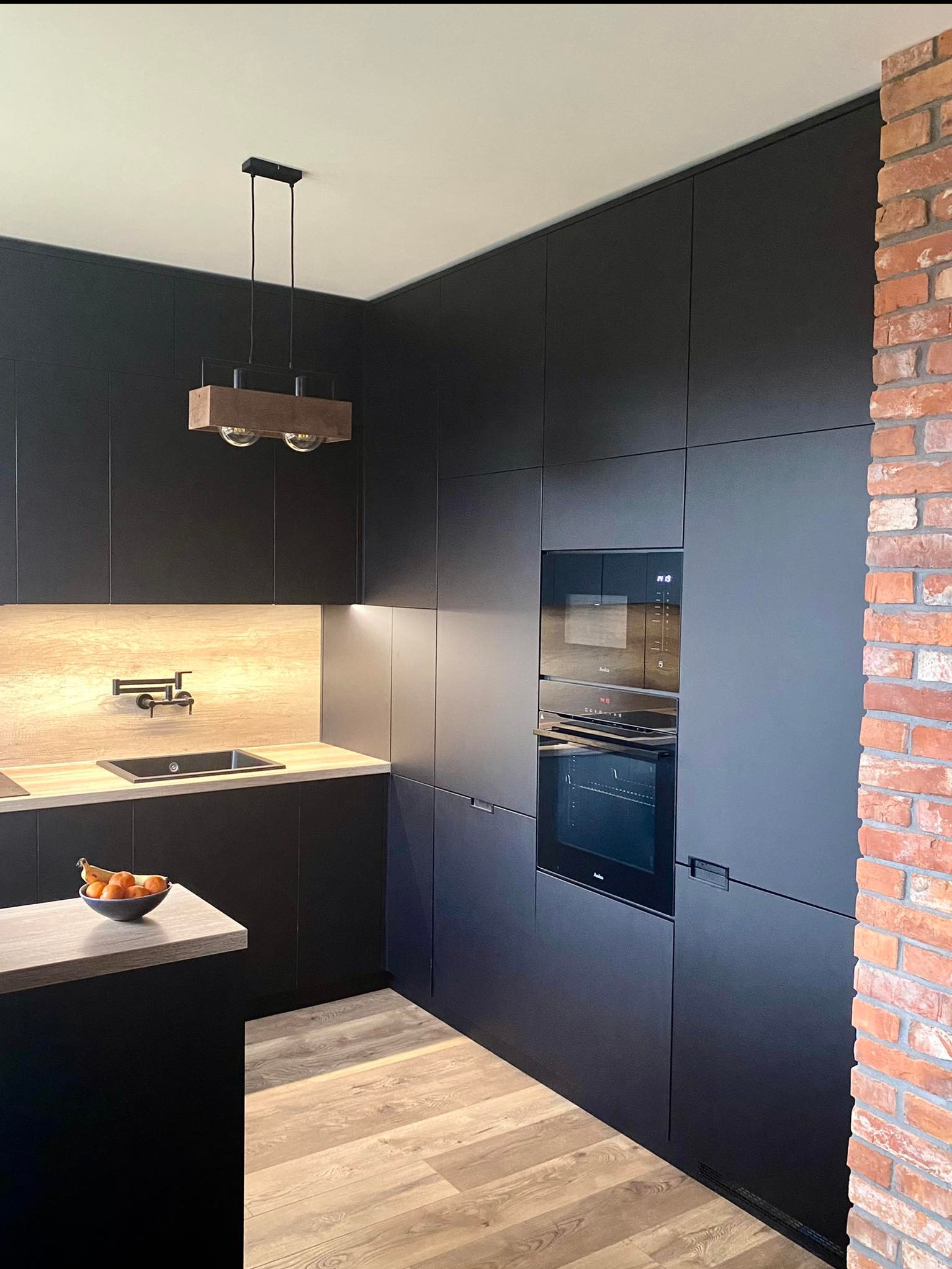Nowoczesna kuchnia z czarnymi, matowymi szafkami, drewnianym blatem i oświetleniem punktowym pod szafkami, widoczny fragment ściany z czerwonej cegły oraz wisząca lampa z drewnianym elementem...