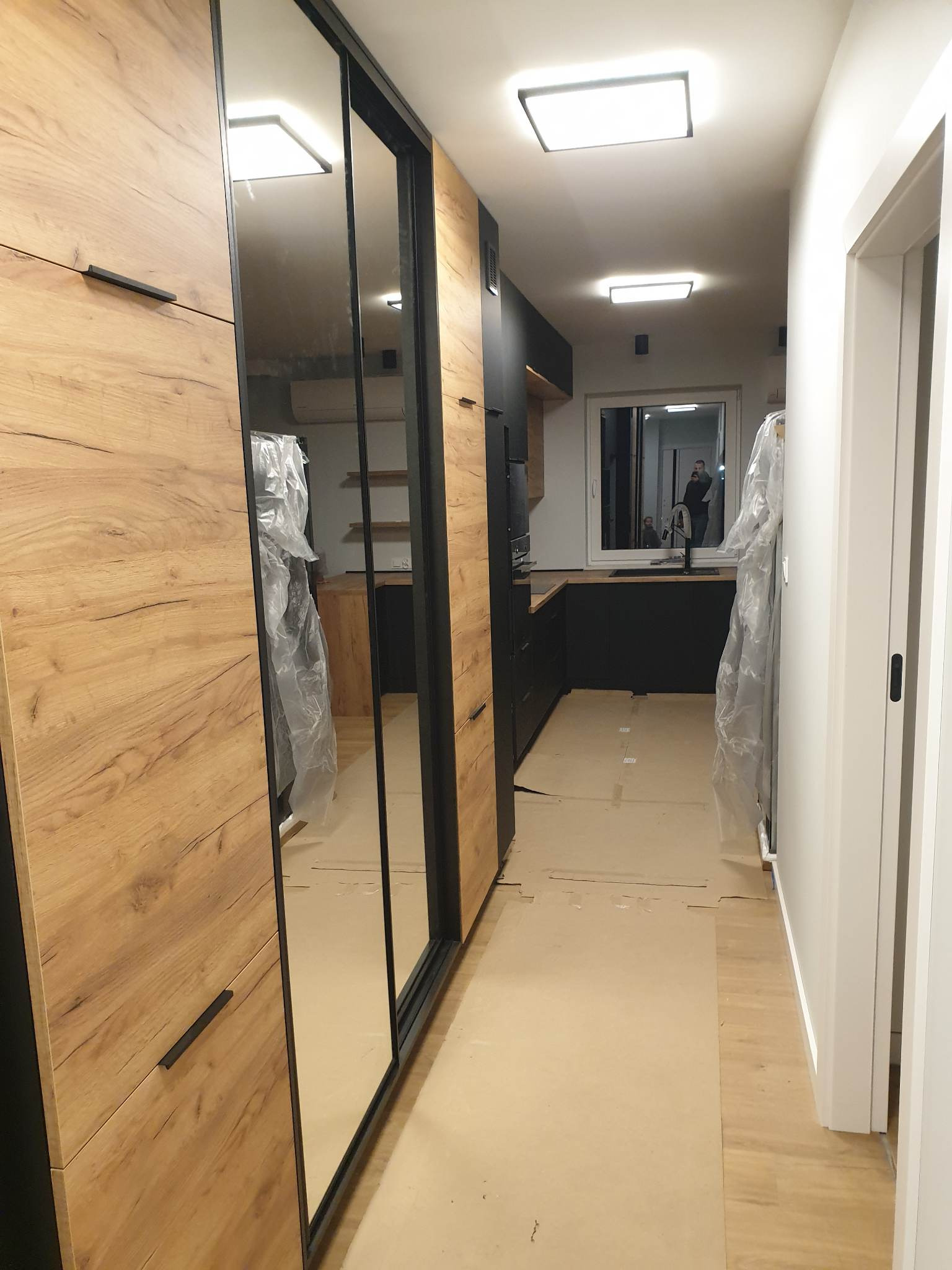Korytarz w trakcie wykańczania z zabudowaną szafą z lustrami i drewnianymi frontami, widok na czarną kuchnię z oknem.