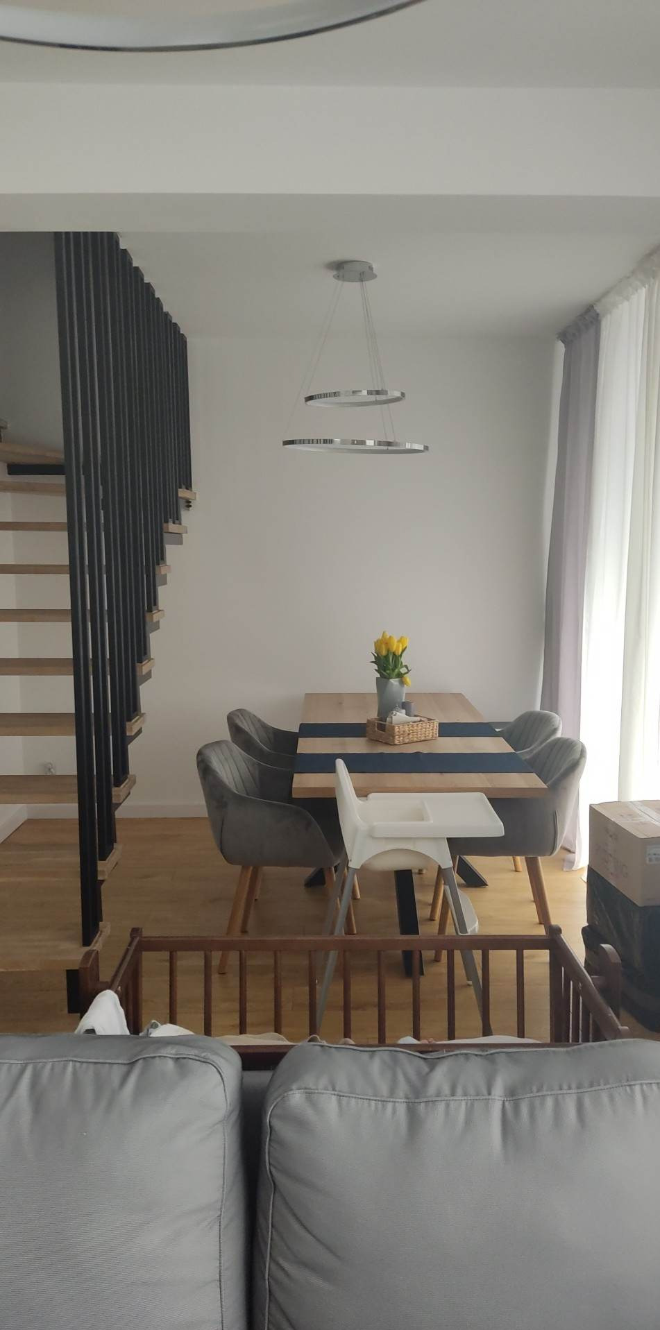 Jasna jadalnia z drewnianym stołem, szarymi krzesłami, białym krzesełkiem do karmienia i dekoracyjnym oświetleniem, widok na drewniane schody z czarną balustradą.