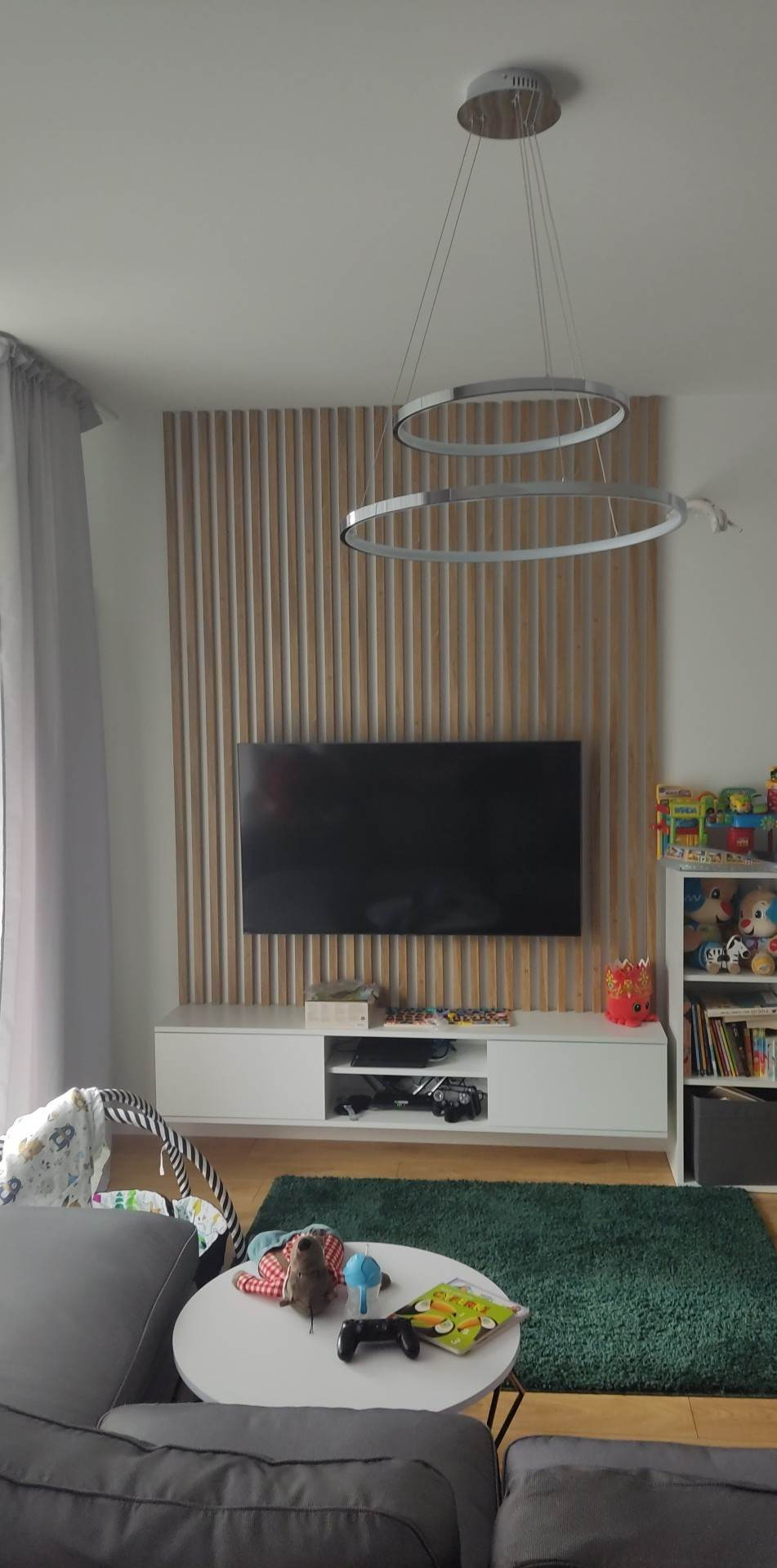 Telewizor na ścianie z drewnianą okładziną, pod nim biała szafka RTV, nad nim dwu-ringowa lampa, obok regał z zabawkami i książkami, w tle szara zasłona i fragment sofy.