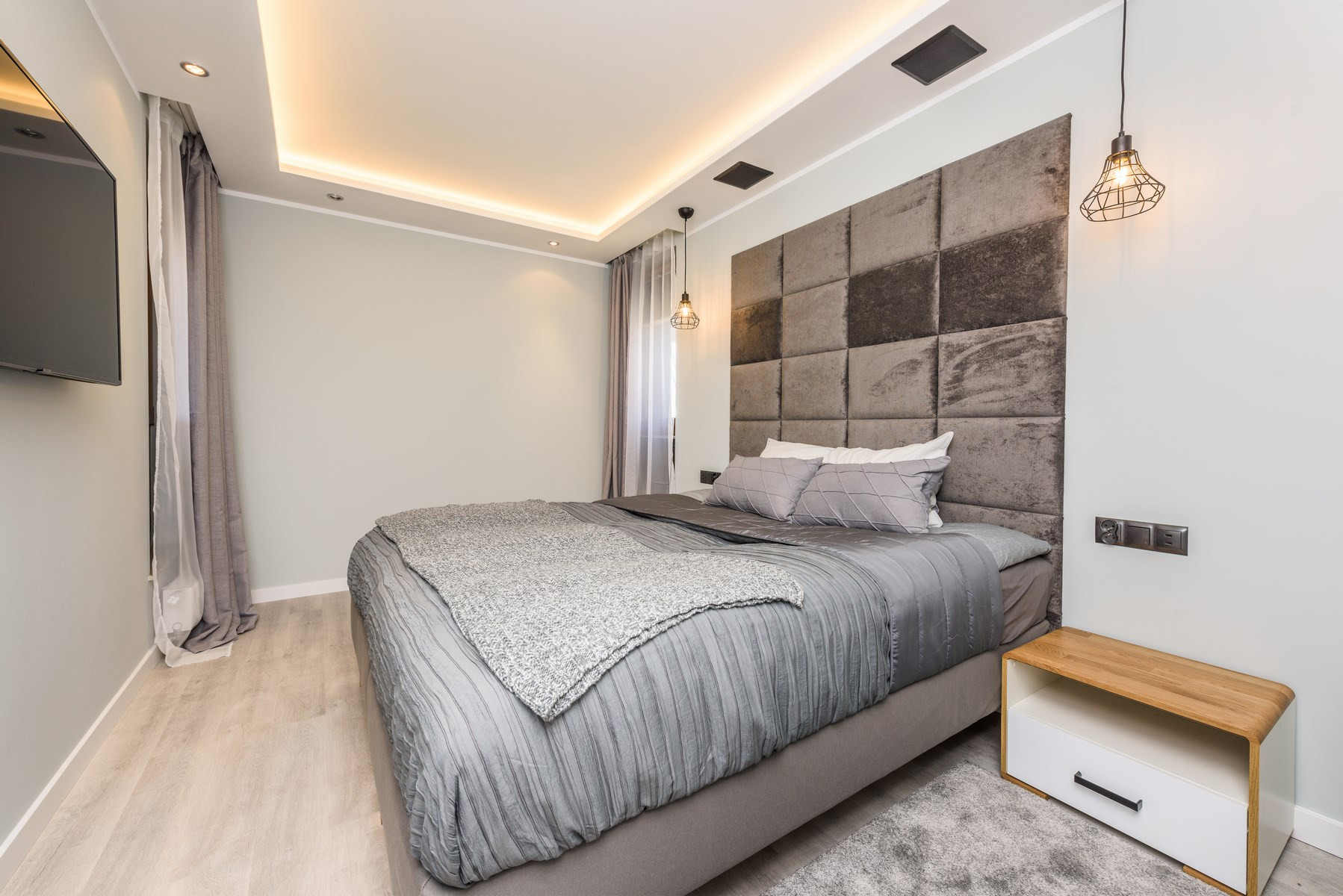 Nowoczesna sypialnia w odcieniach szarości z tapicerowanym zagłówkiem w kwadraty, wiszącymi lampami w stylu industrialnym i drewnianą szafką nocną z białą szufladą.