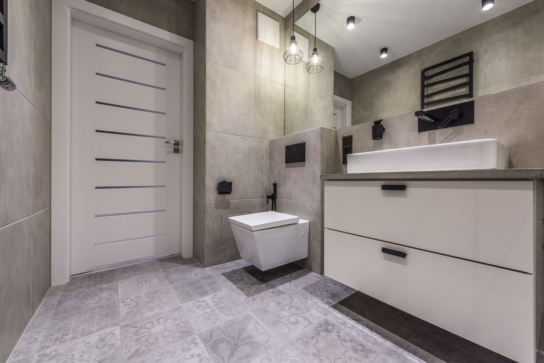 Nowoczesna łazienka w szarościach z białą wiszącą toaletą, umywalką nablatową i wzorzystą podłogą, oświetlona lampami wiszącymi w stylu industrialnym i punktowymi halogenami.