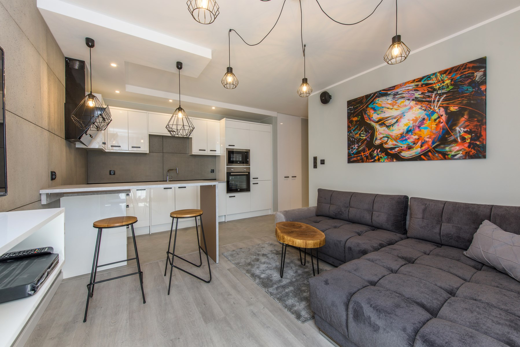 Nowoczesne wnętrze: otwarta kuchnia z białymi szafkami i szarym backsplashiem, wyspa z drewnianymi stołkami barowymi, szara sofa narożna i abstrakcyjny obraz z profilem twarzy na ścianie...