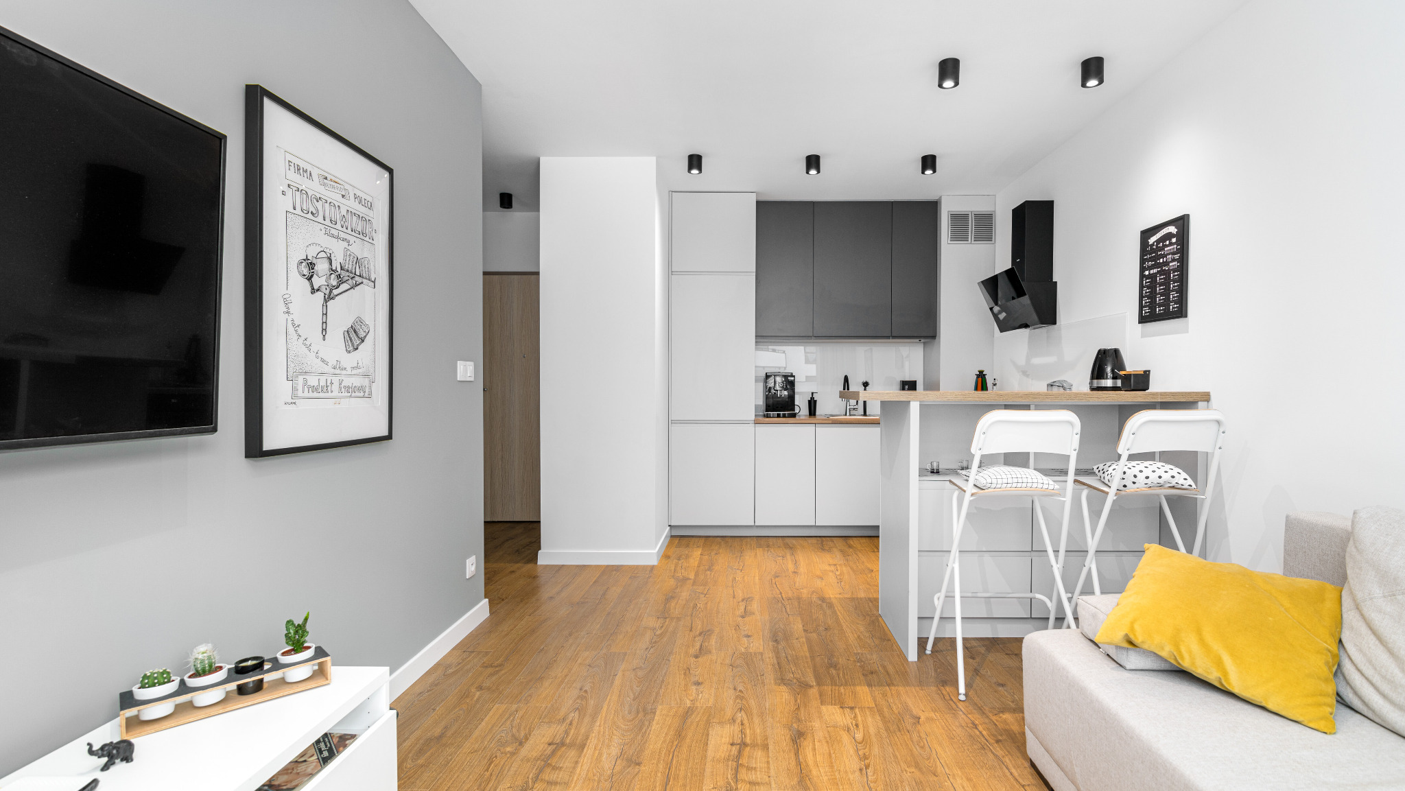 Jasne, nowoczesne wnętrze mieszkania z drewnianą podłogą, aneksem kuchennym z szarymi i białymi szafkami, barkiem z białymi krzesłami, szarą ścianą z czarnym telewizorem i plakatem w ramie.