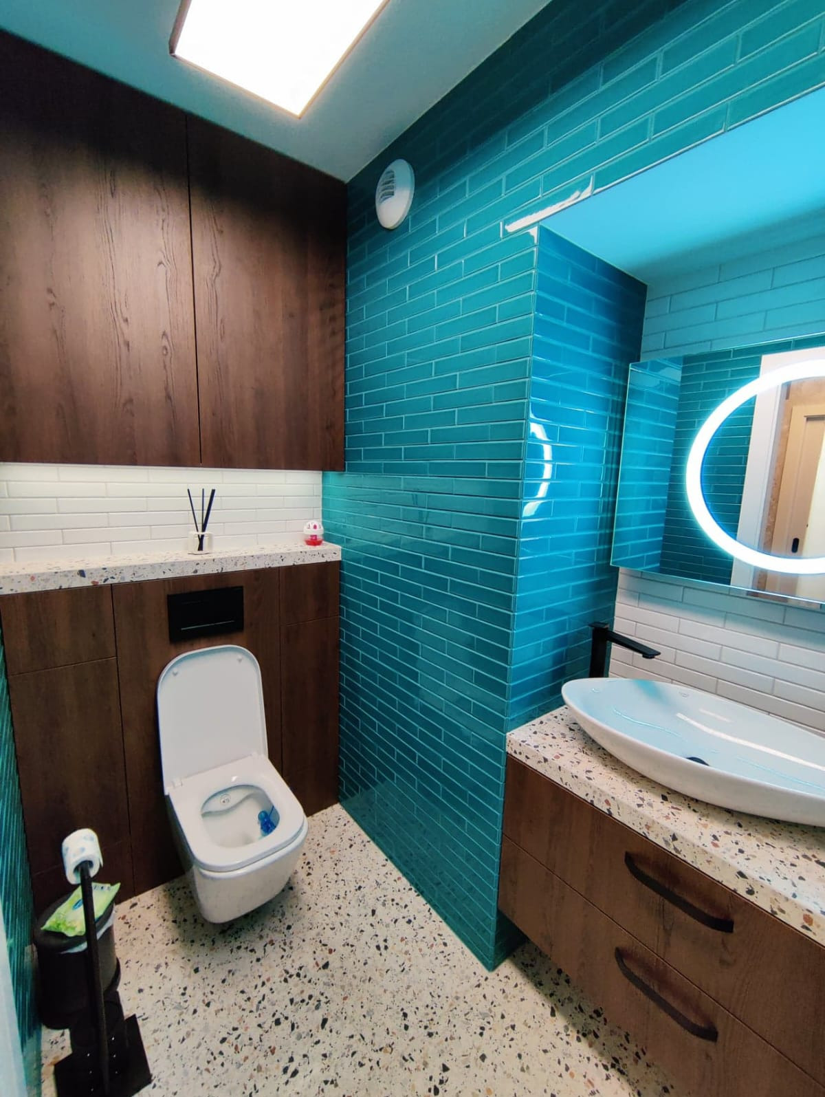 Nowoczesna łazienka z turkusowymi płytkami na ścianie, wiszącą toaletą i szafkami z ciemnego drewna, uzupełniona okrągłym lustrem z podświetleniem LED i podłogą z lastryko.