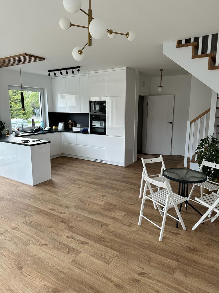 Jasna kuchnia z białymi, błyszczącymi szafkami i czarnym blatem w Gdańsku, widok na stół z krzesłami i schody z drewnianymi stopniami.