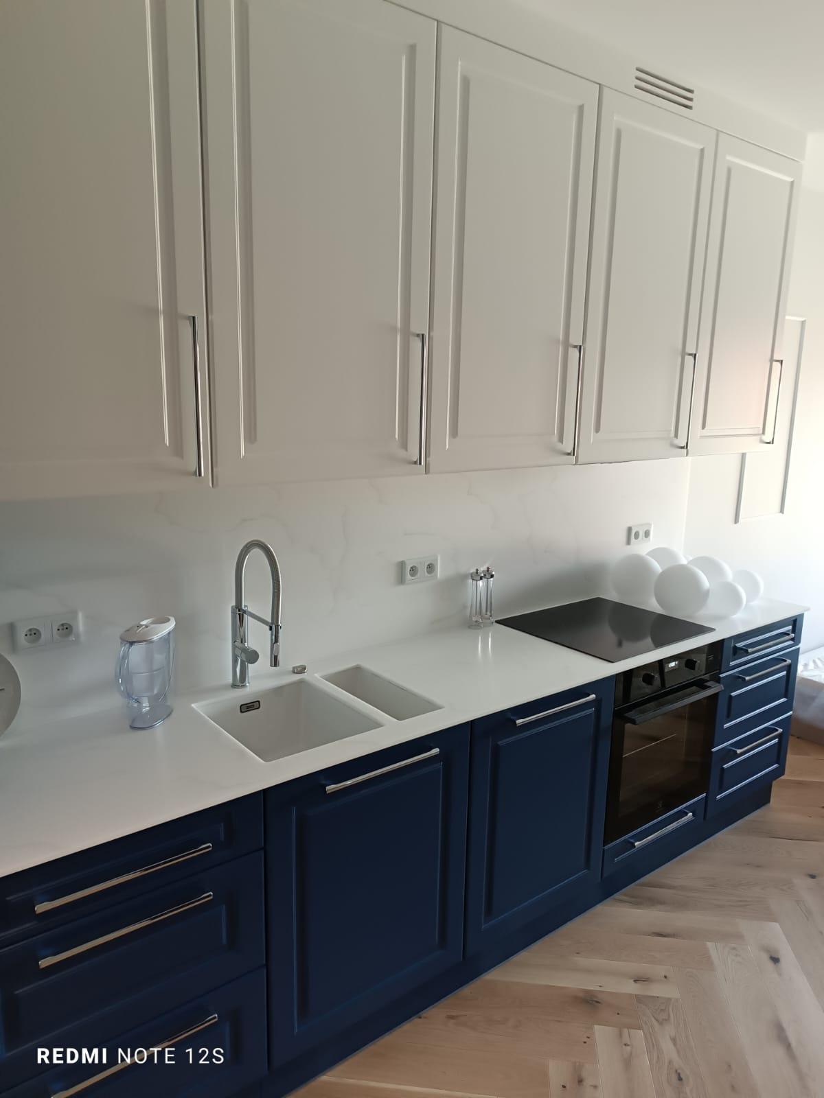 Nowoczesna kuchnia z białymi szafkami górnymi i granatowymi dolnymi, biały blat z marmurowym wzorem, zlewozmywak dwukomorowy i płyta indukcyjna, drewniana podłoga ułożona w jodełkę.