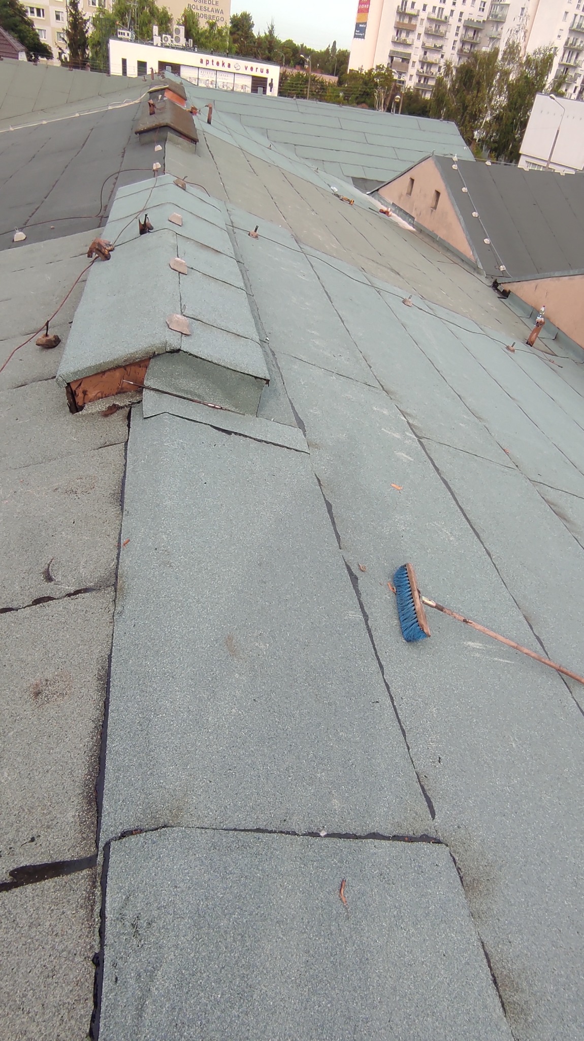 Fragment dachu pokrytego papą w Poznaniu, z widocznym kominem i szczotką dekarską leżącą na powierzchni. Widoczne także budynki w tle.