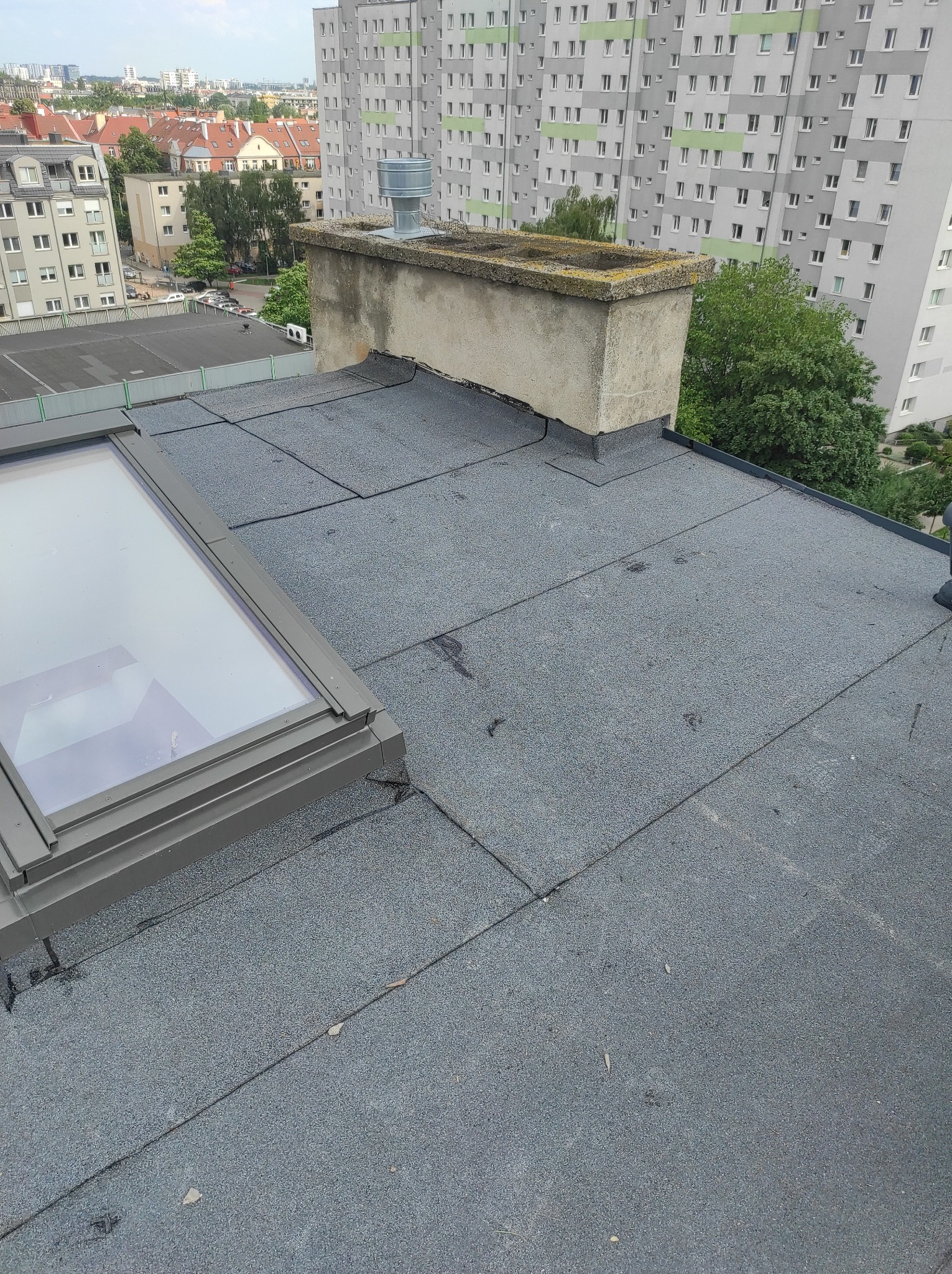 Widok z góry na płaski dach pokryty papą z widocznym oknem dachowym, kominem z lekkim porostem mchu i blokami mieszkalnymi w tle.