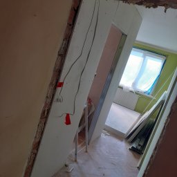 Chrystian śipek - Montaż płyt kartonowo-gipsowych w trakcie remontu, widoczne kable elektryczne przytwierdzone czerwoną taśmą, surowa cegła w narożniku, podłoga z parkietu w jodełkę.