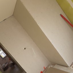 Chrystian śipek - Montaż płyt gipsowo-kartonowych na poddaszu, widoczny poziomica, otwór na instalację elektryczną i fragment okna w tle.