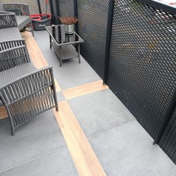 Chrystian śipek - Wykończony balkon z szarymi płytkami, drewnopodobnymi wstawkami, meblami ogrodowymi z technorattanu i ażurową balustradą.
